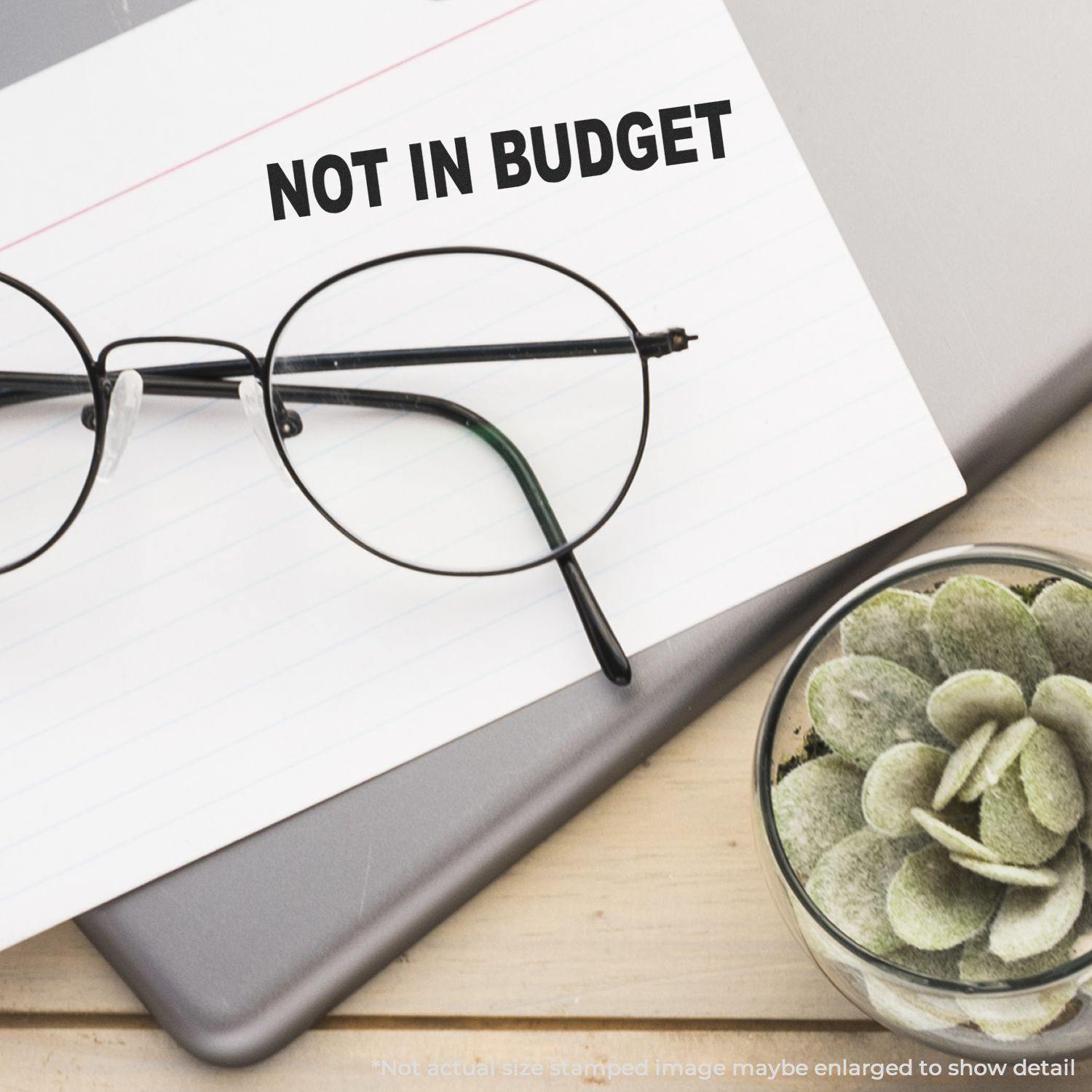 A Large Not In Budget Rubber Stamp is used on a white card placed on a laptop, with glasses and a small succulent plant nearby.