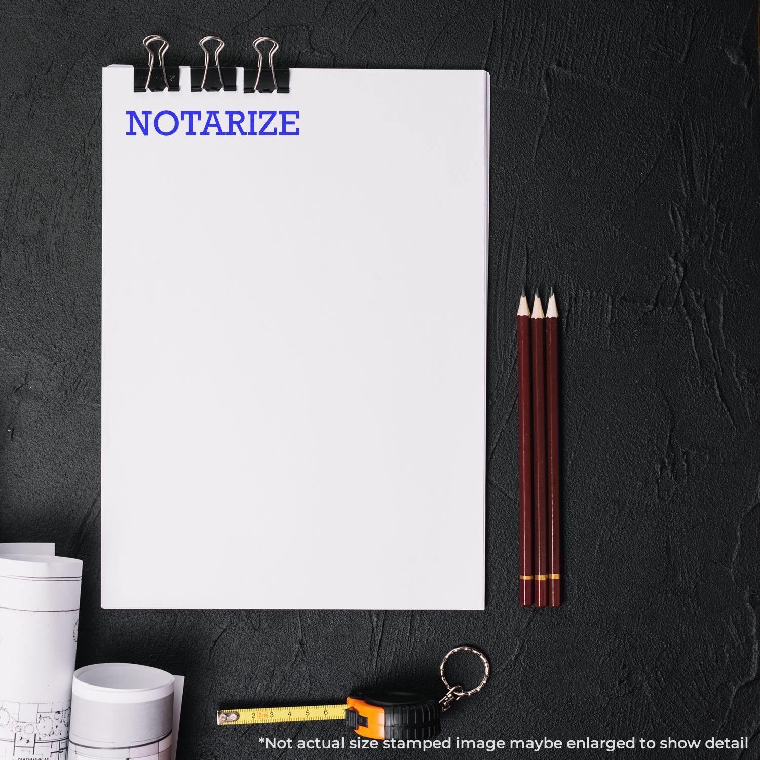 A Large Pre-Inked Notarize Stamp is used on a white sheet of paper, with pencils, a tape measure, and blueprints on a black surface.