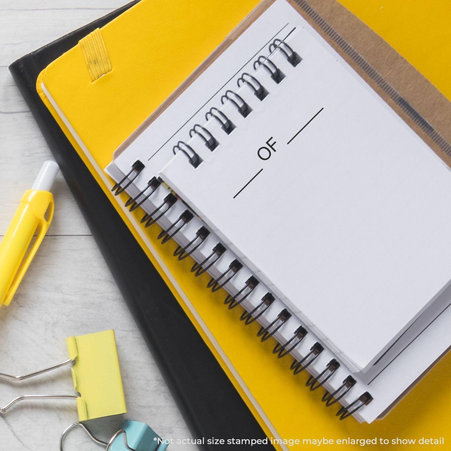 Large Pre-Inked OF Stamp on a white notepad, surrounded by yellow and black notebooks, a pen, and binder clips on a desk.