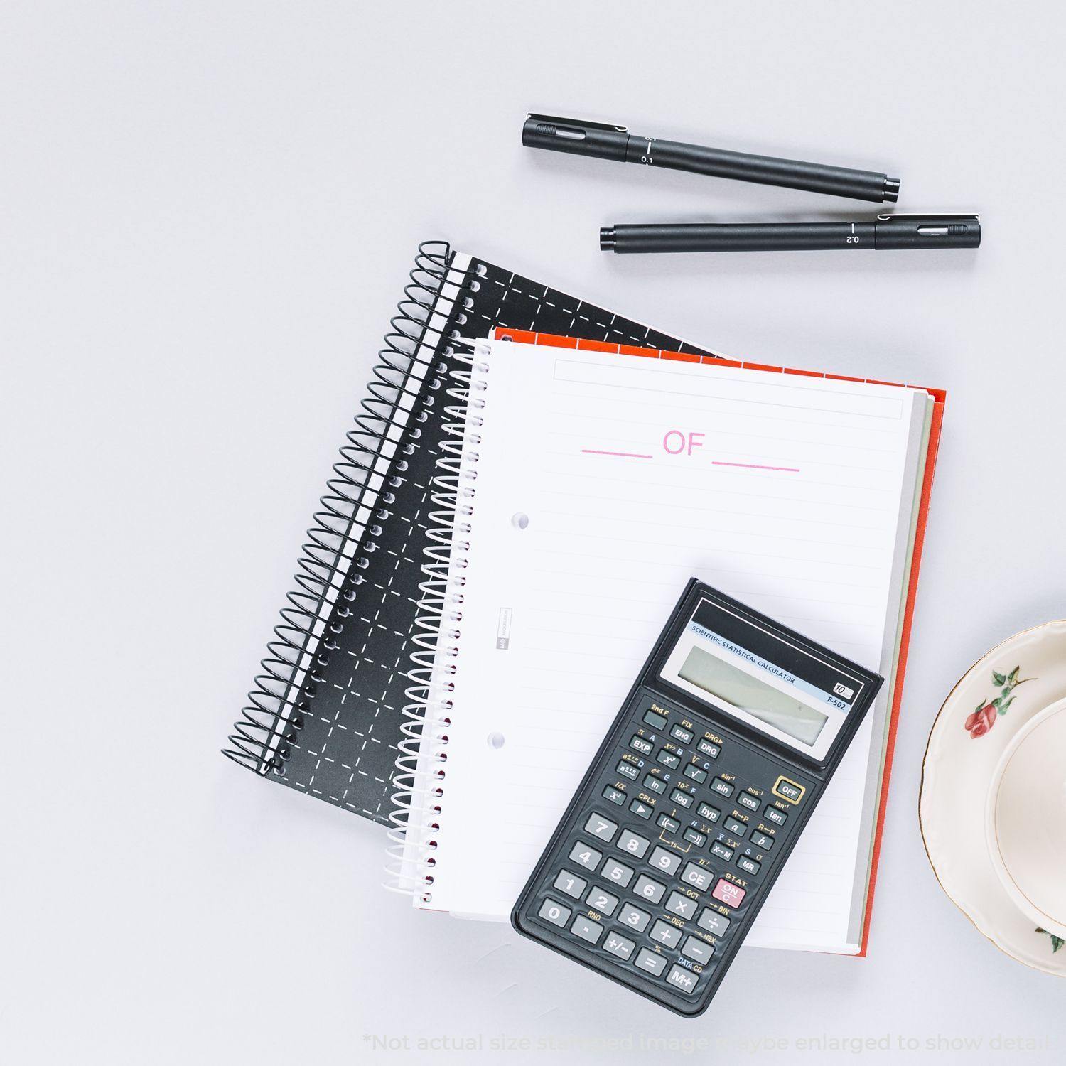 Large Self Inking Of Stamp in use on a white notebook, surrounded by a calculator, two pens, and other stationery on a desk.