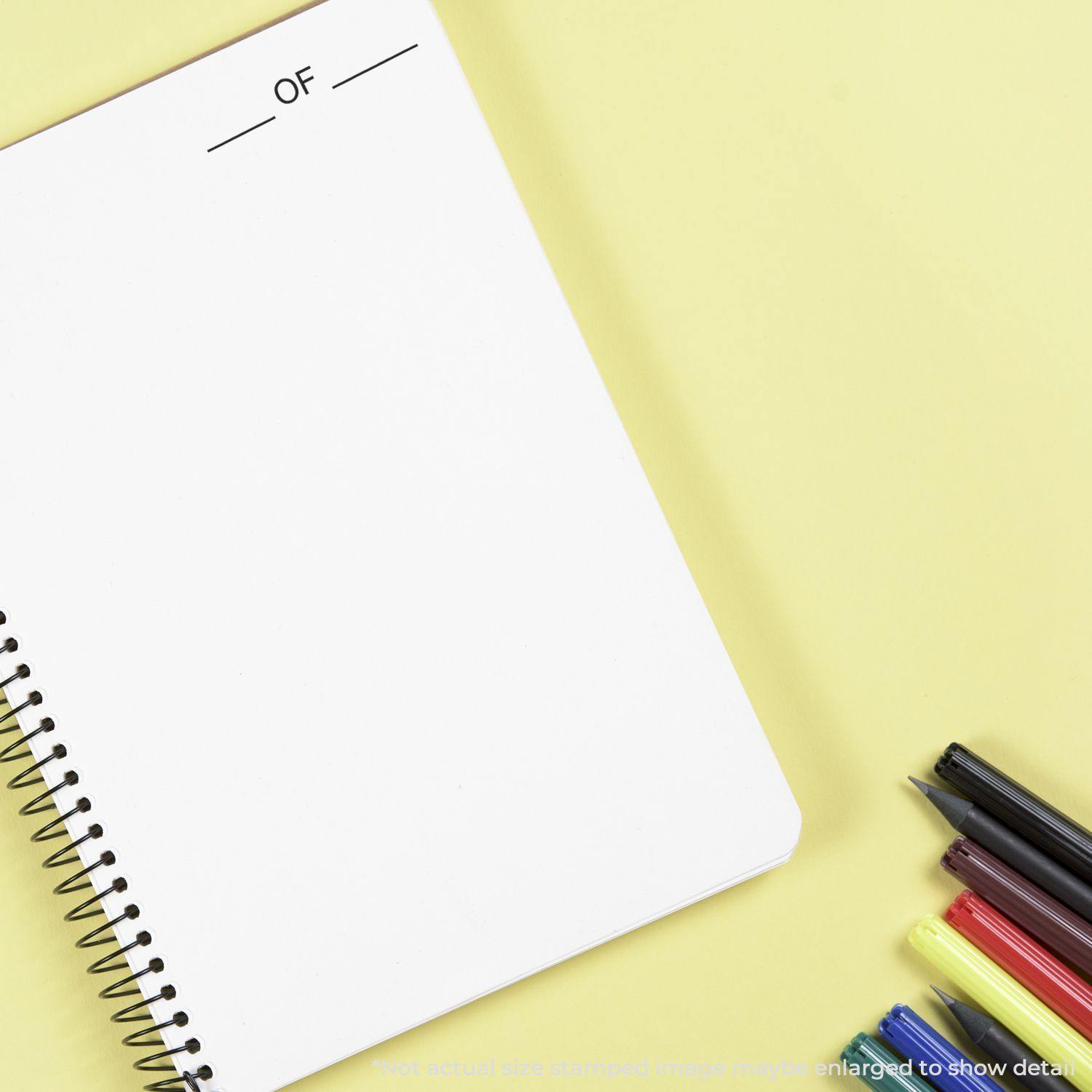 Large Pre-Inked OF Stamp used on a spiral notebook, with pens and highlighters placed beside it on a yellow background.