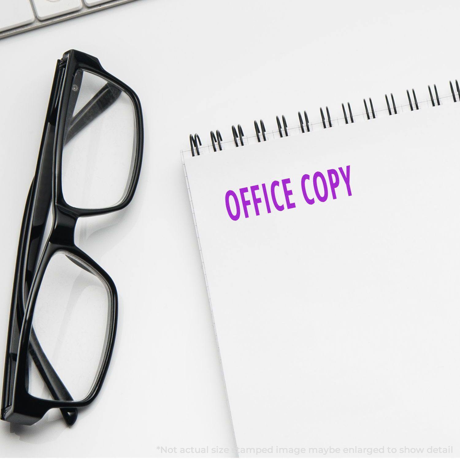 Slim Pre-Inked Office Copy Stamp in purple ink on a white notepad, next to black eyeglasses on a white desk.