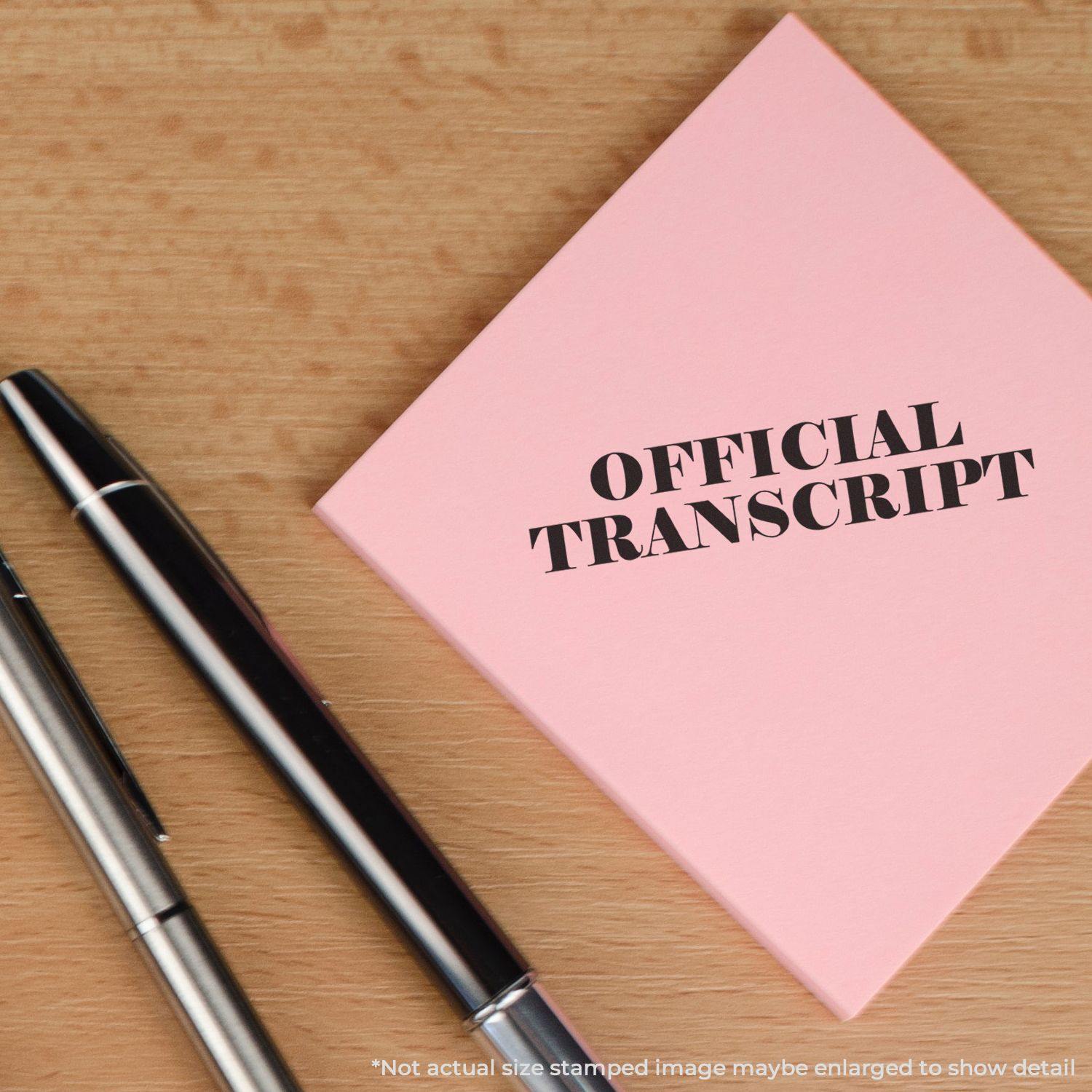 Large Pre-Inked Official Transcript Stamp used on a pink paper next to two pens on a wooden surface.