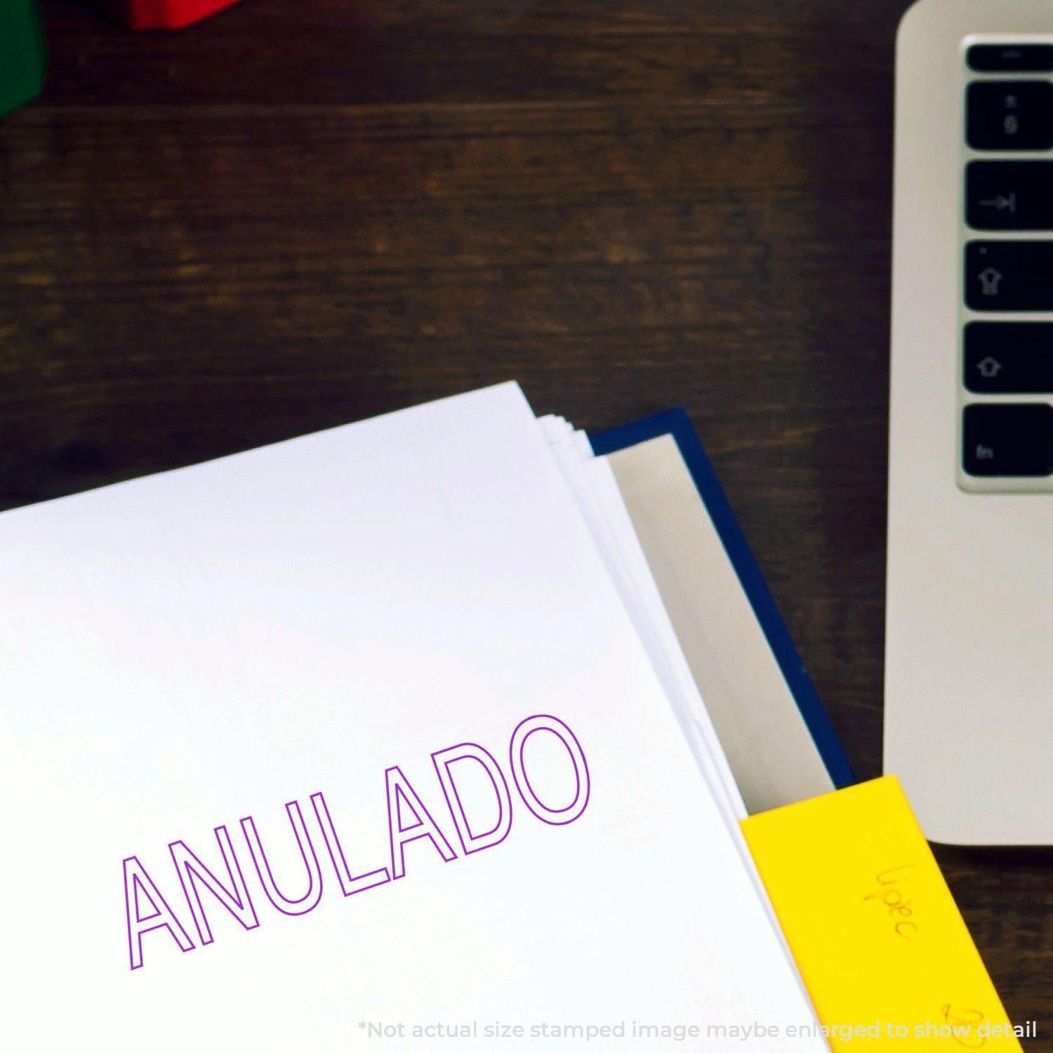 Large Outline Anulado Rubber Stamp used on a white paper, placed on a wooden desk next to a laptop and a yellow sticky note.