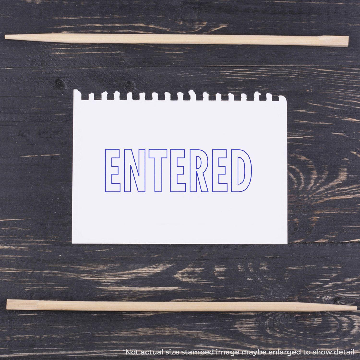 Large Entered With Outline Rubber Stamp used on a white notepad, displaying the word ENTERED in blue outline letters on a dark wooden surface.