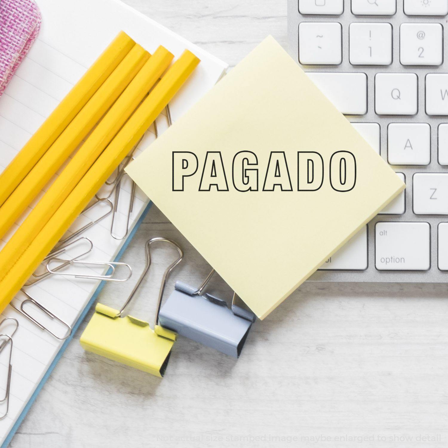 Slim Pre-Inked Outline Pagado Stamp on a sticky note beside a keyboard, pencils, and paper clips on a desk.