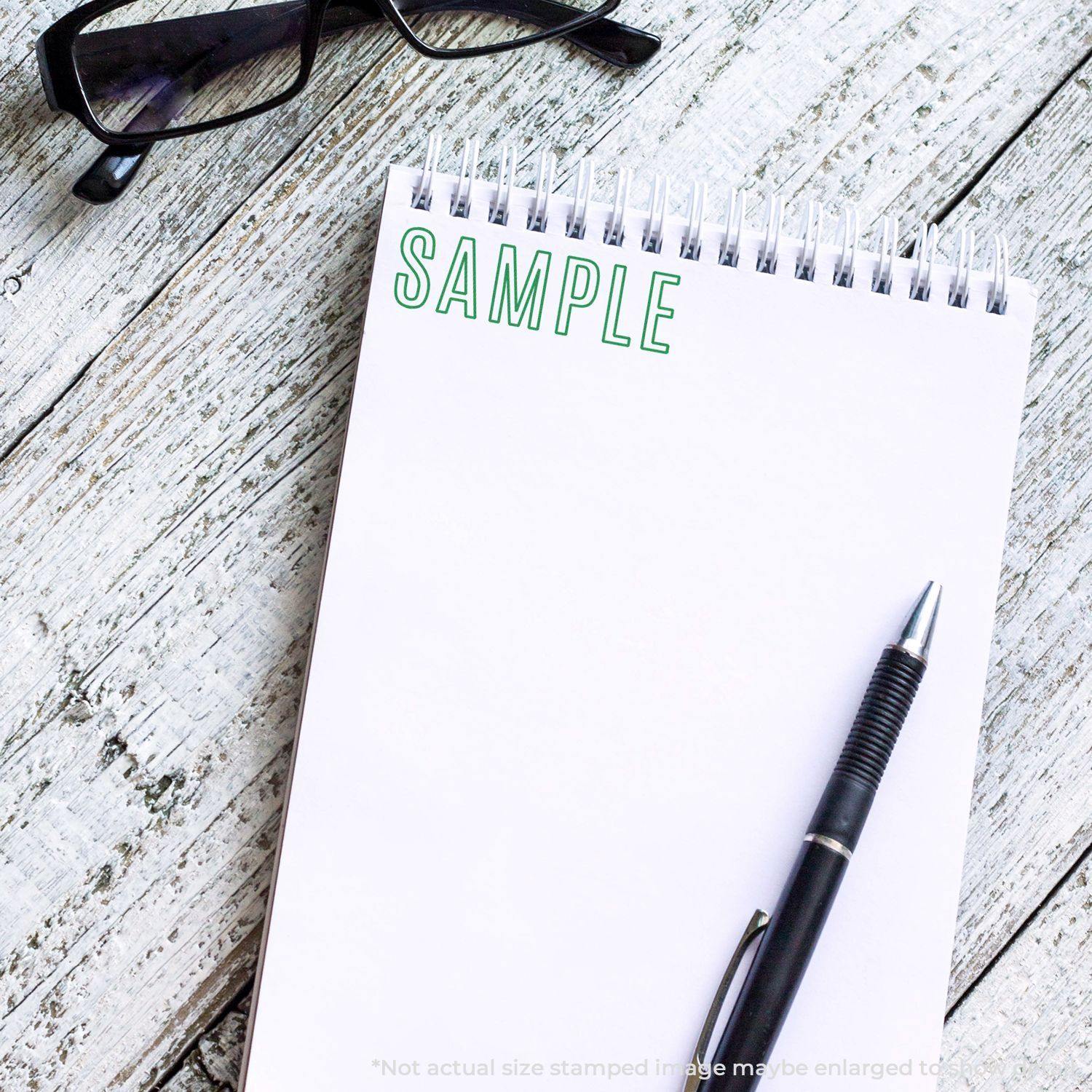 Large Outline Sample Rubber Stamp used on a white notepad with a pen and glasses placed on a wooden surface.