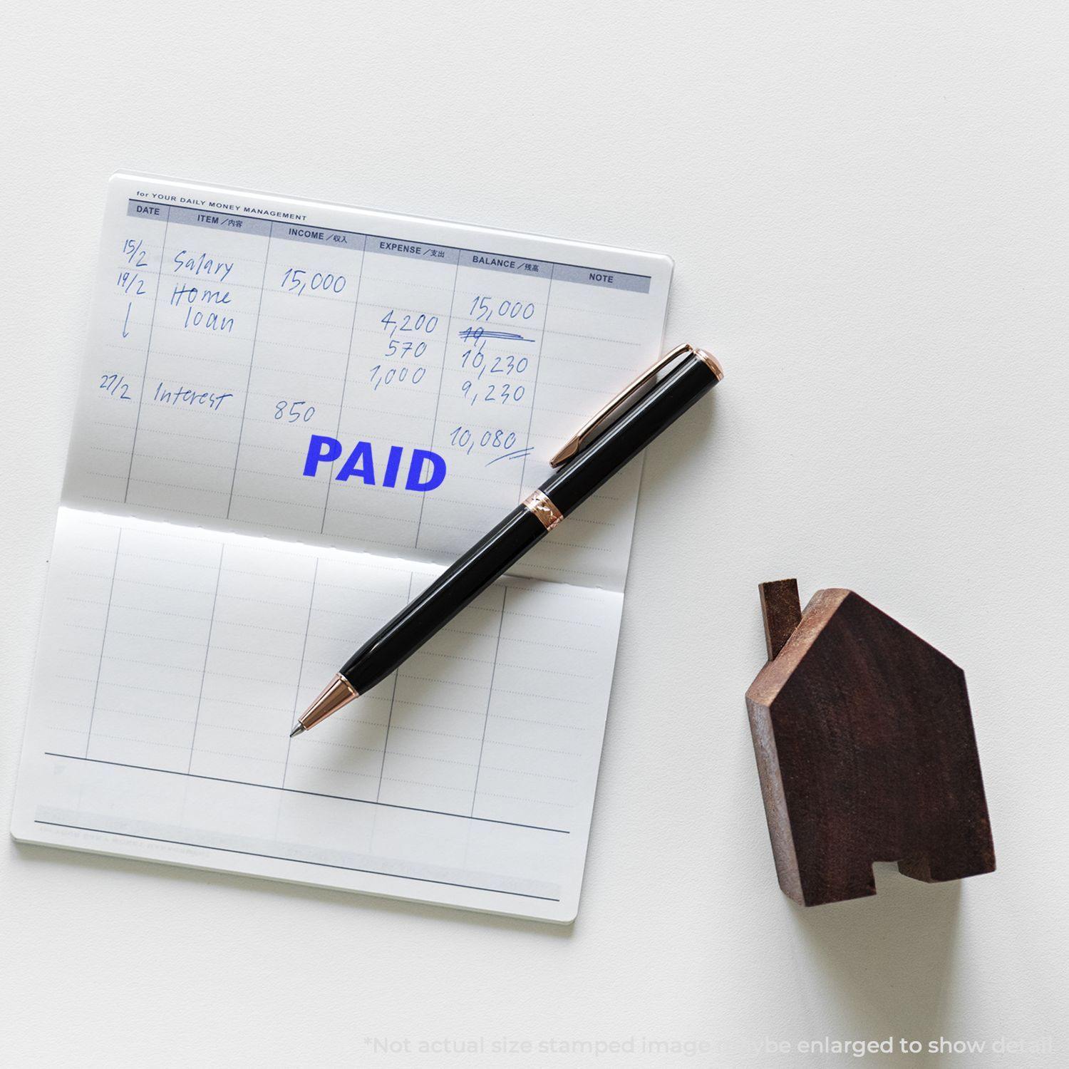 A Large Self Inking Paid Stamp is used on a financial ledger, with a pen and a wooden house-shaped object placed nearby.