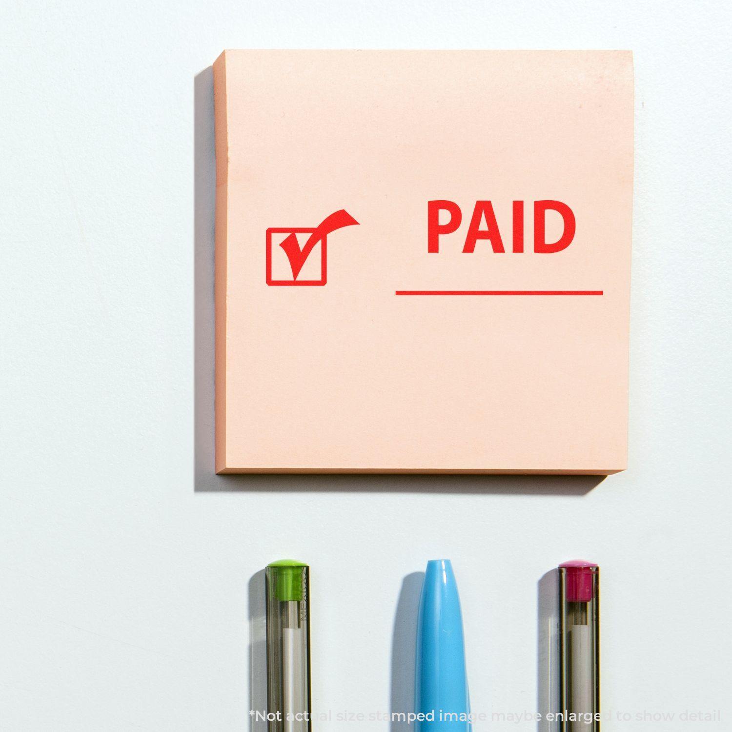 Paid with Checkmark Rubber Stamp on a pink sticky note, with three pens below it on a white surface.