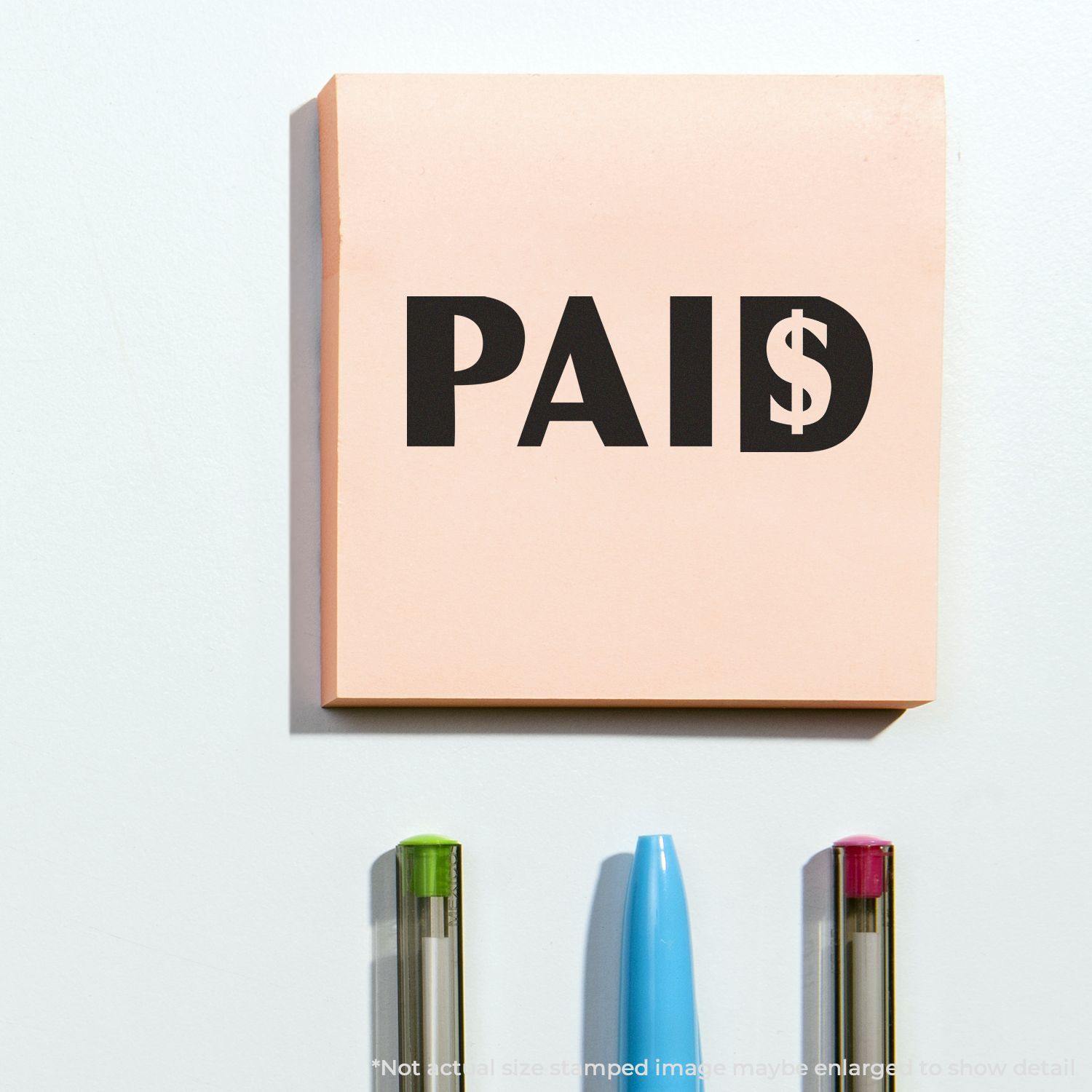 Paid with Dollar Sign Rubber Stamp on a pink sticky note, with three pens below it on a white surface.