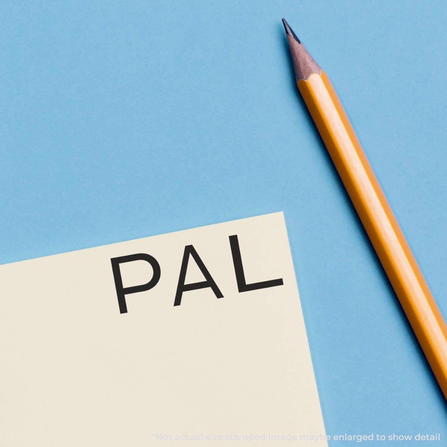 A Slim Pre-Inked PAL Stamp imprint on a yellow paper next to a sharpened yellow pencil on a blue background.