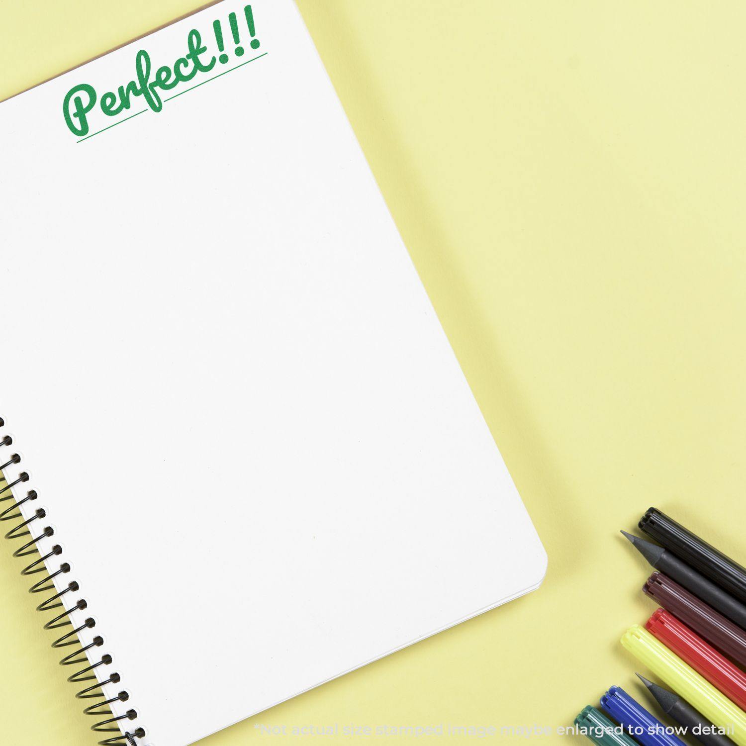 Large Pre-Inked Perfect Stamp in green ink on a white spiral notebook, with colorful pens and markers on a yellow background.