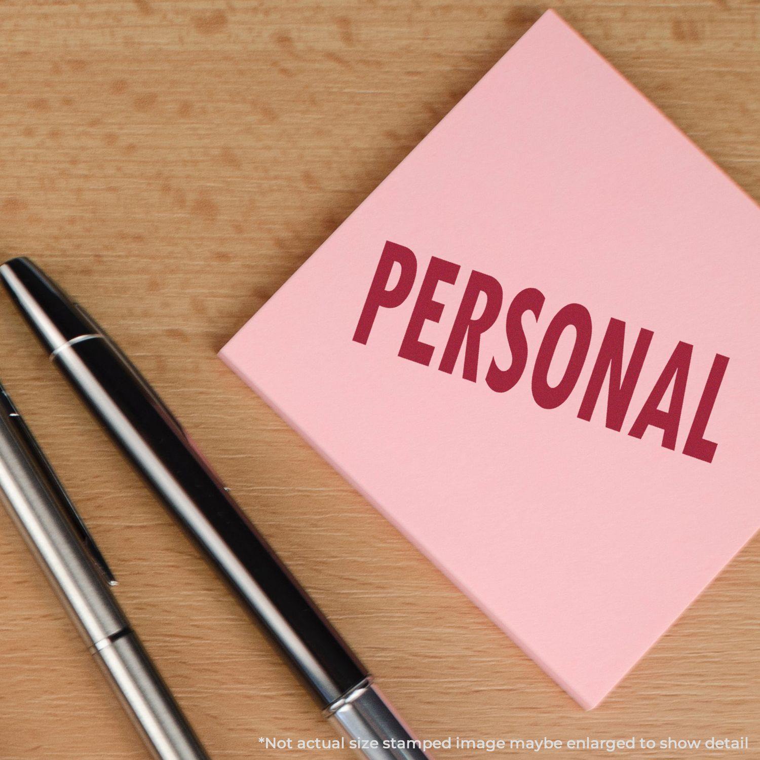 Large Personal Rubber Stamp imprint on a pink sticky note with the word PERSONAL next to two pens on a wooden surface.