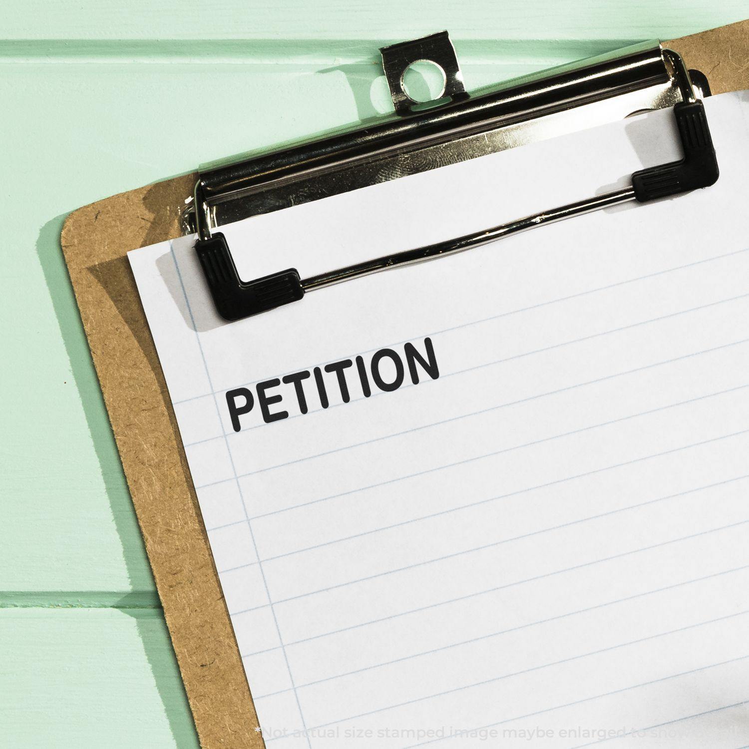 A clipboard with a sheet of paper stamped PETITION using a Slim Pre-Inked Petition Stamp, placed on a light green surface.