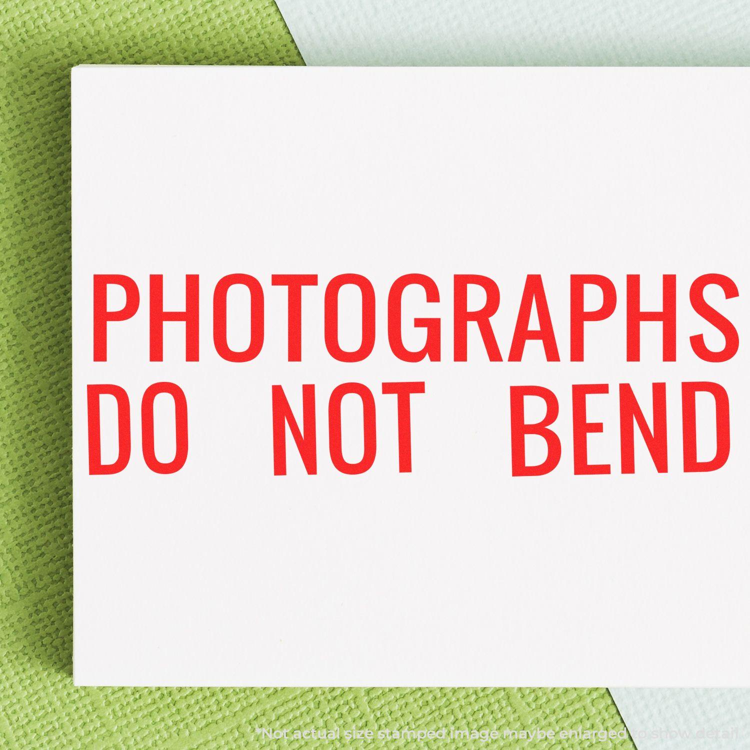 Photographs Do Not Bend rubber stamp in red ink on white paper, placed on a green and white textured background.