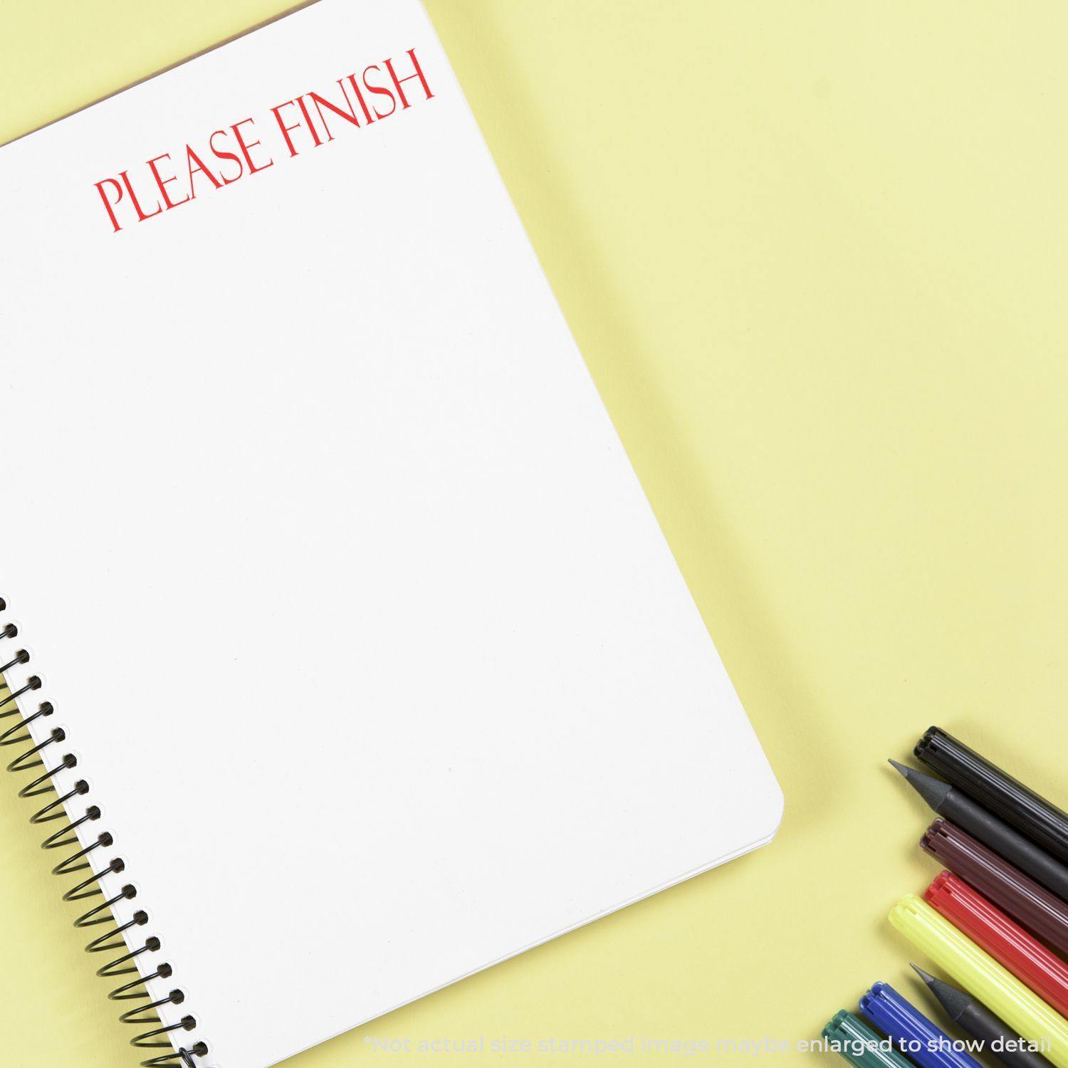 A notebook with PLEASE FINISH stamped in red using a Slim Pre-Inked Please Finish Stamp, next to colorful pens on a yellow background.