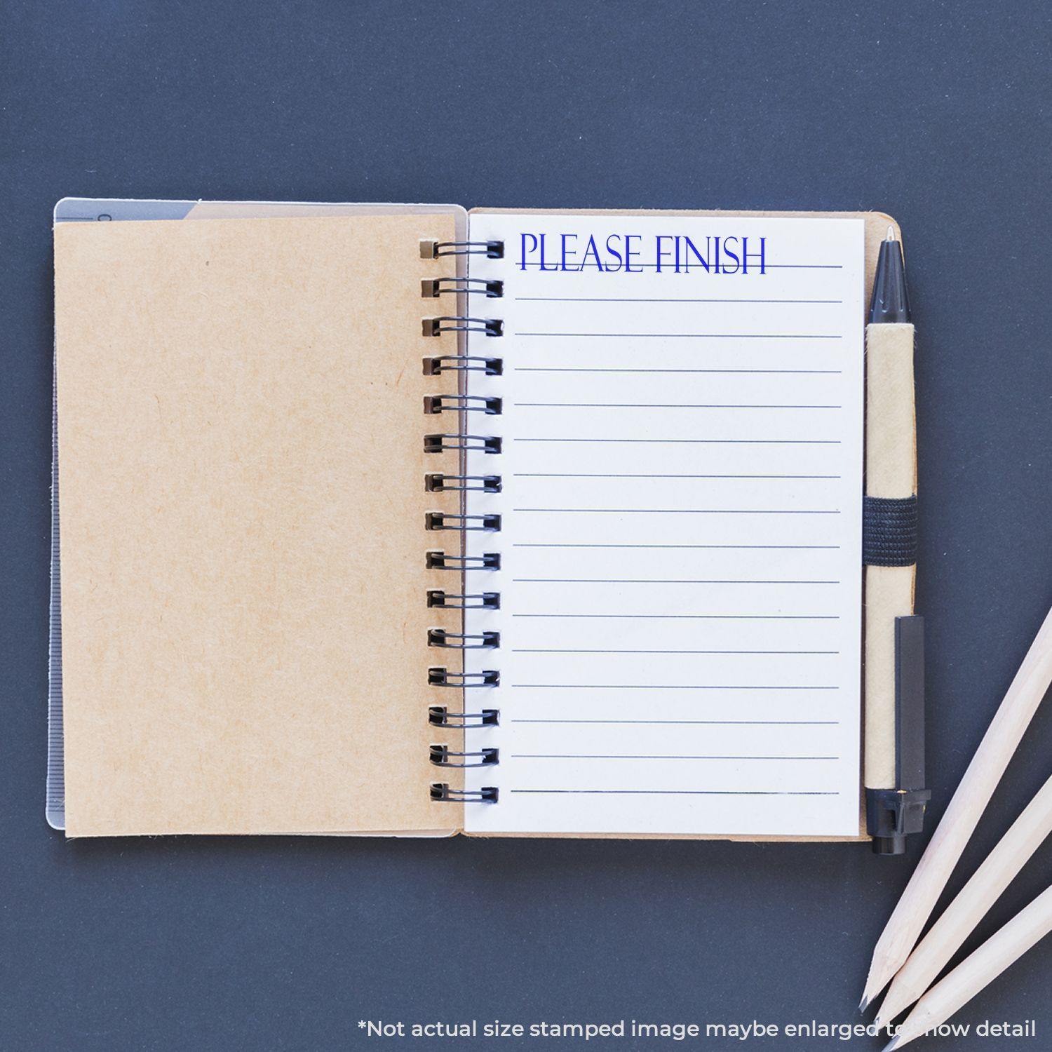 A notebook with a Please Finish rubber stamp mark on a lined page, surrounded by a pen and pencils on a dark surface.