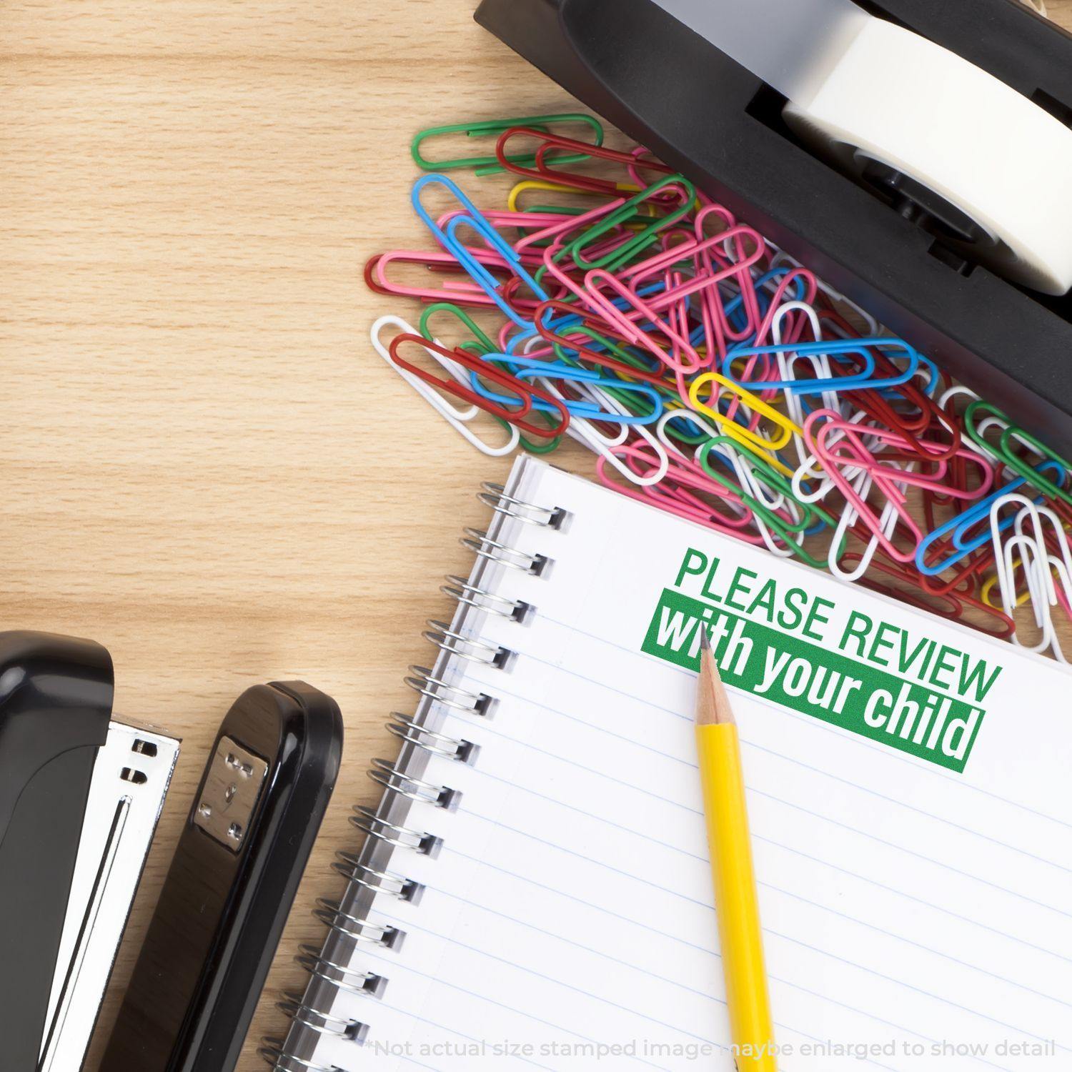 A desk with a stapler, tape dispenser, colorful paperclips, and a notebook stamped with Large Please Review with your child Rubber Stamp in green.