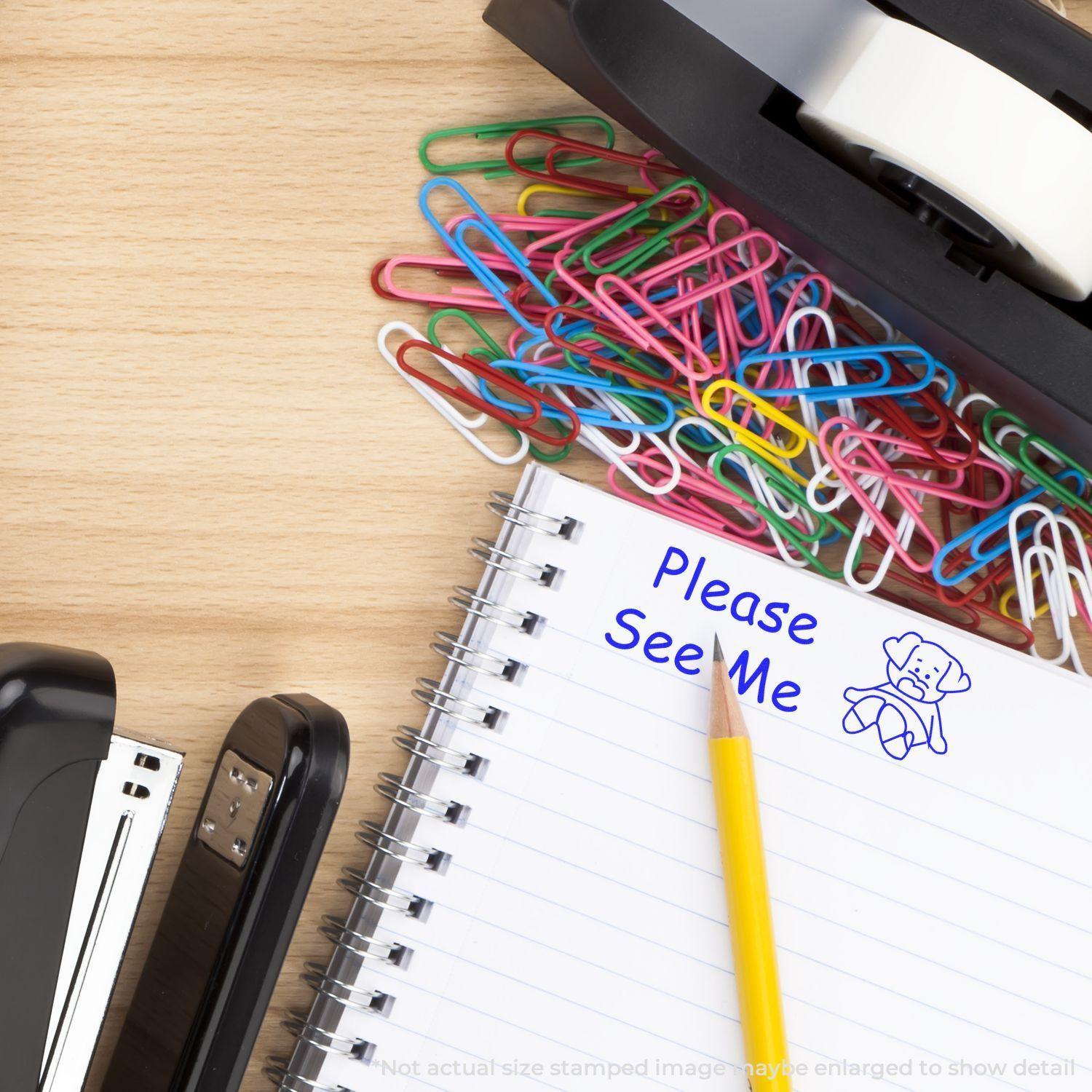 A desk with a Large Pre-Inked Please See Me Stamp impression on a notebook, surrounded by colorful paperclips, a stapler, and a tape dispenser.