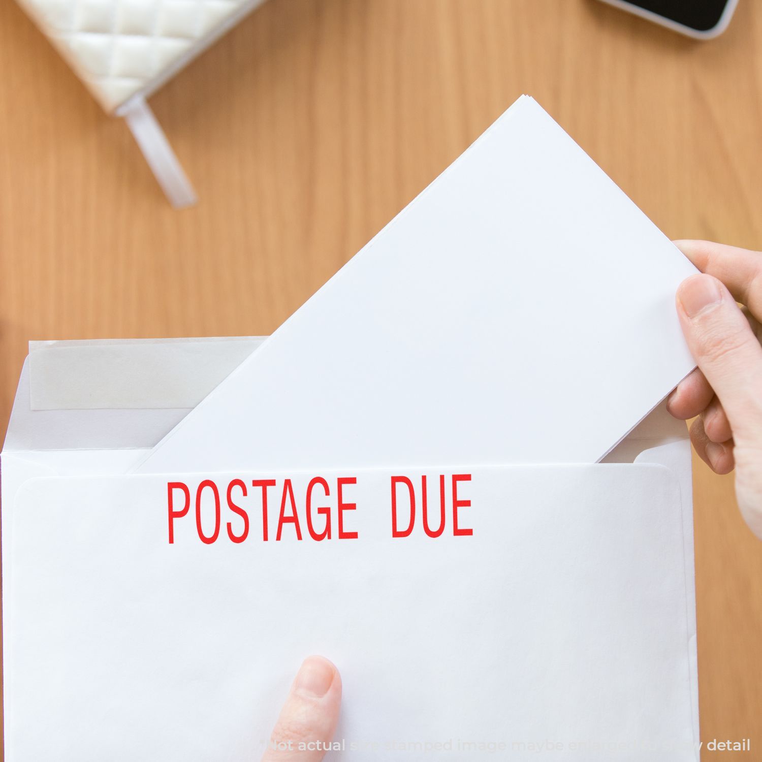 Person holding an envelope stamped with POSTAGE DUE in red using a Self Inking Postage Due Stamp on a wooden desk.