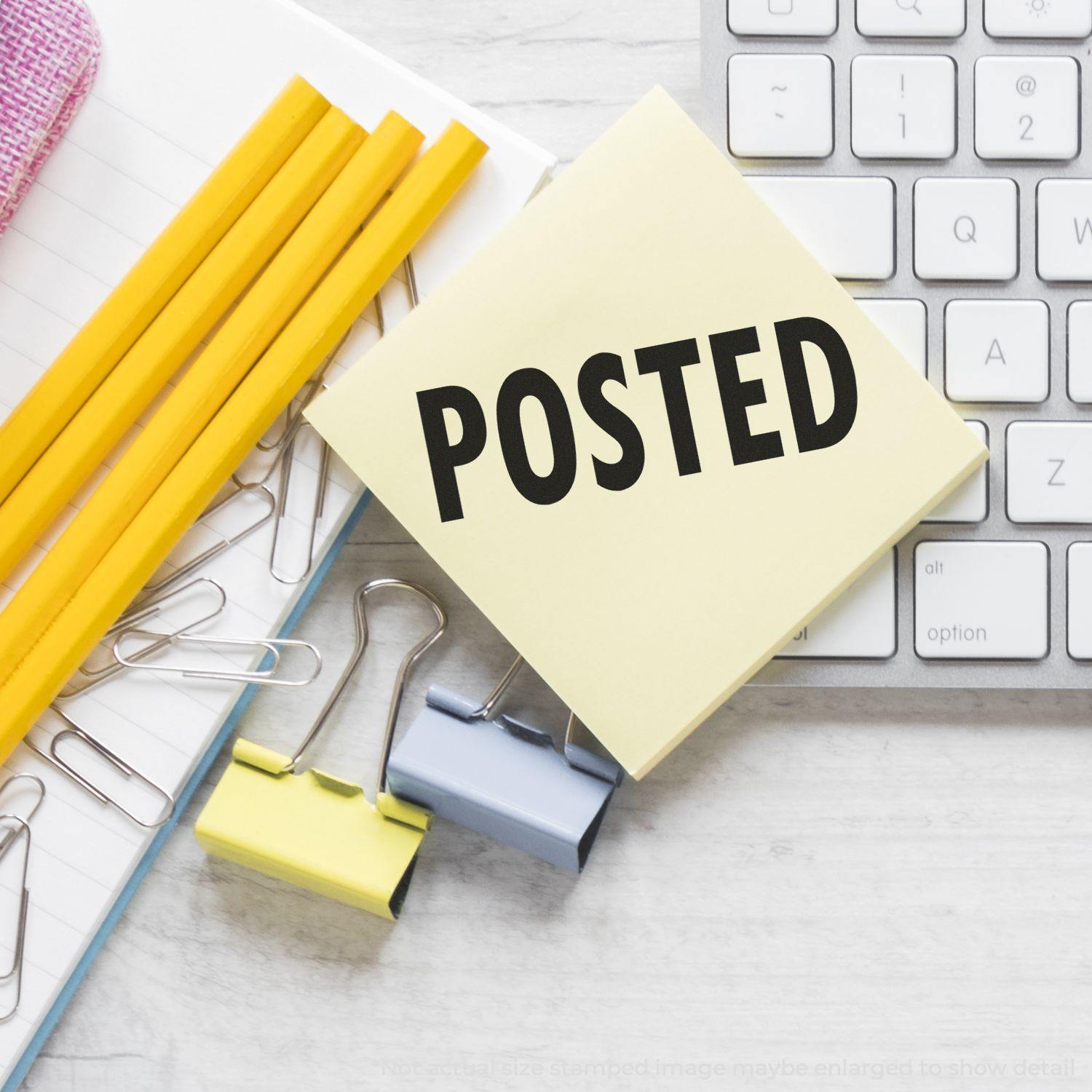 Large Posted Rubber Stamp on a yellow sticky note beside a keyboard, pencils, paper clips, and binder clips on a desk.