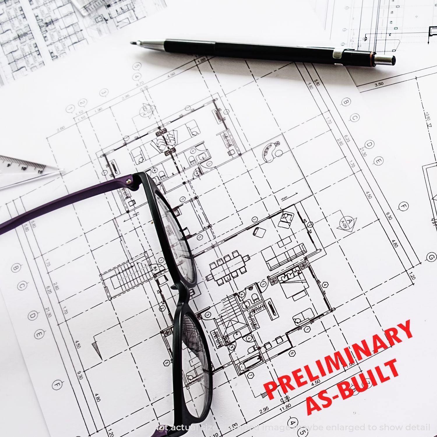 Architectural plans with a Self Inking Preliminary As Built Stamp in red ink, alongside a pen and glasses on a white background.