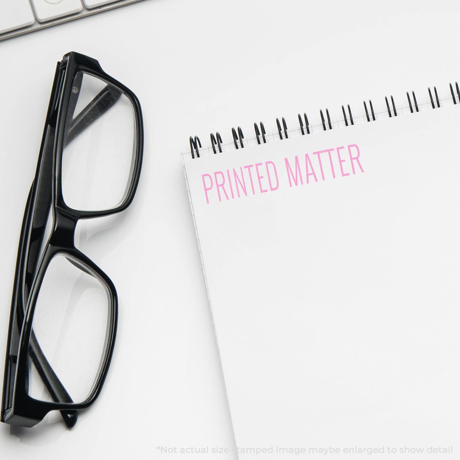 Large Self Inking Printed Matter Stamp in use, stamping PRINTED MATTER in pink on a white notepad, next to black glasses.