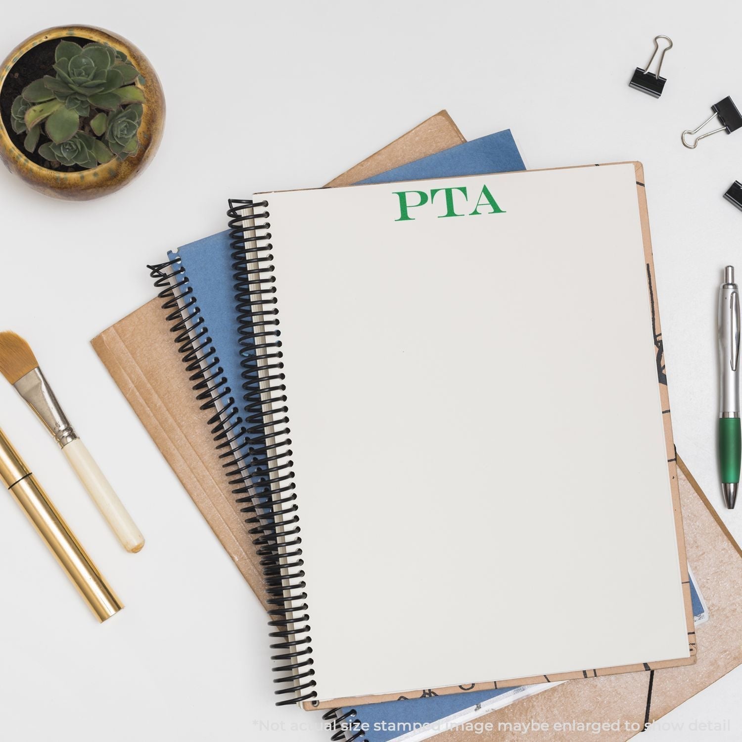 Self Inking PTA Stamp used on a spiral notebook, surrounded by office supplies including a pen, clips, brushes, and a plant.