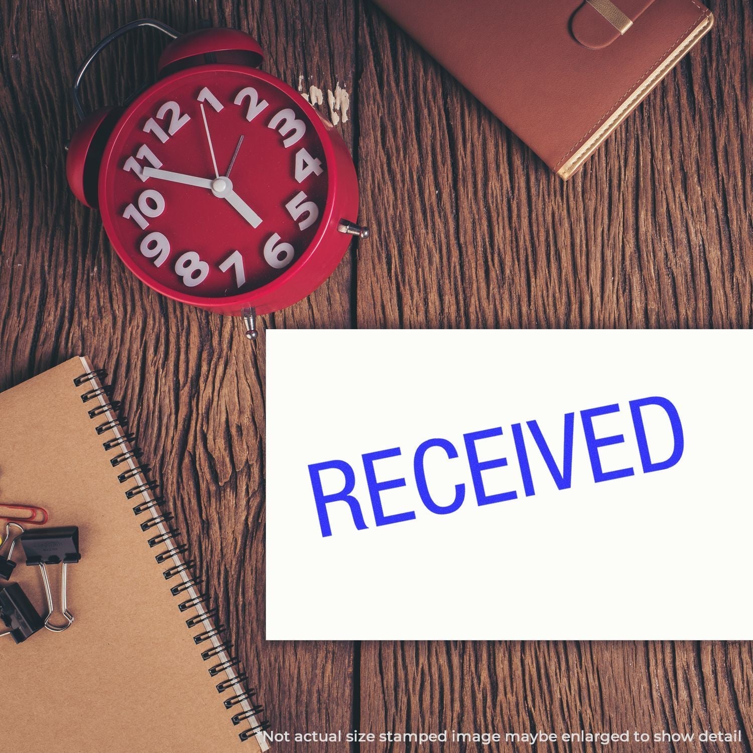 A Self-Inking Received Stamp marks a white paper on a wooden desk with a red clock, notebook, and office supplies nearby.