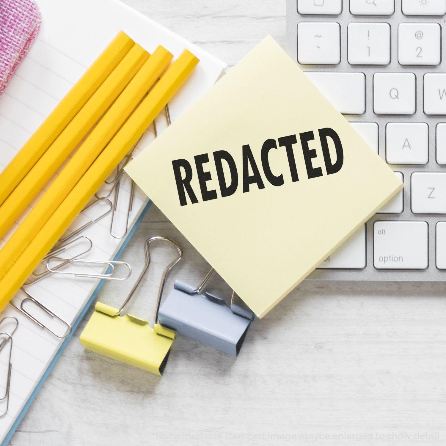 Large Pre-Inked Redacted Stamp on a sticky note beside pencils, paper clips, binder clips, and a keyboard on a desk.