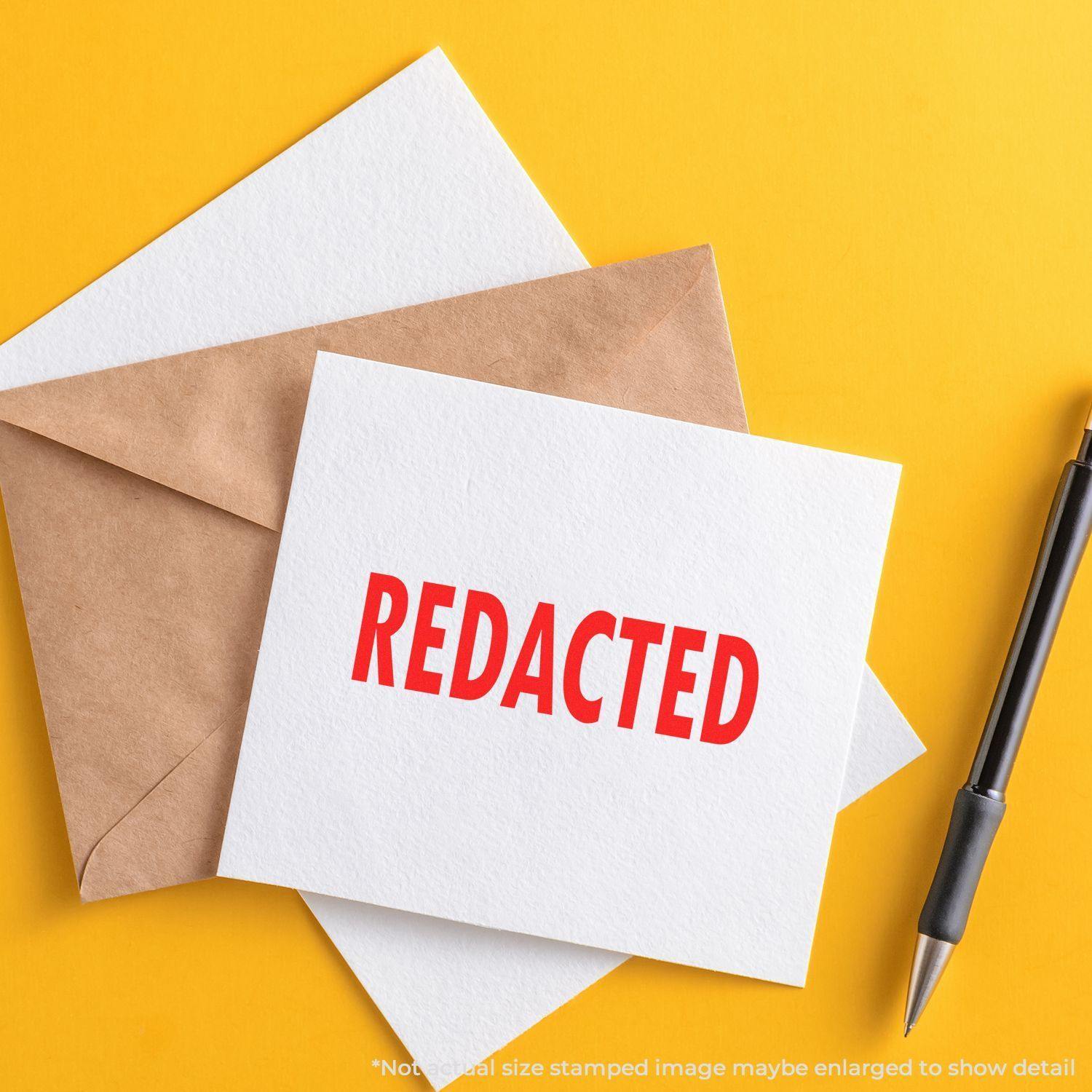 Envelope and card stamped with REDACTED using the Large Pre-Inked Redacted Stamp, placed on a yellow background with a pen nearby.