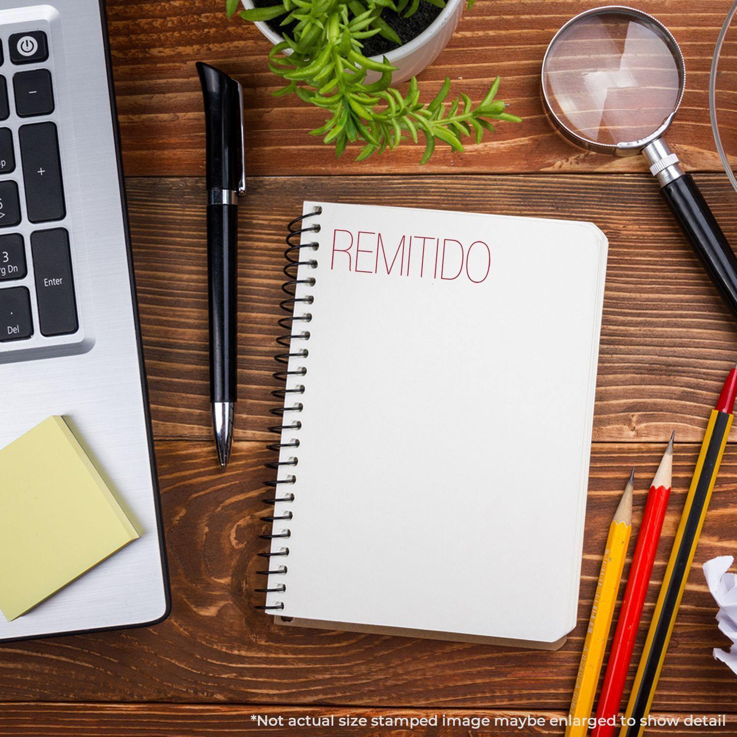 Large Pre-Inked Remitido Stamp used on a notebook, placed on a wooden desk with a laptop, pen, sticky notes, plant, magnifying glass, and pencils.