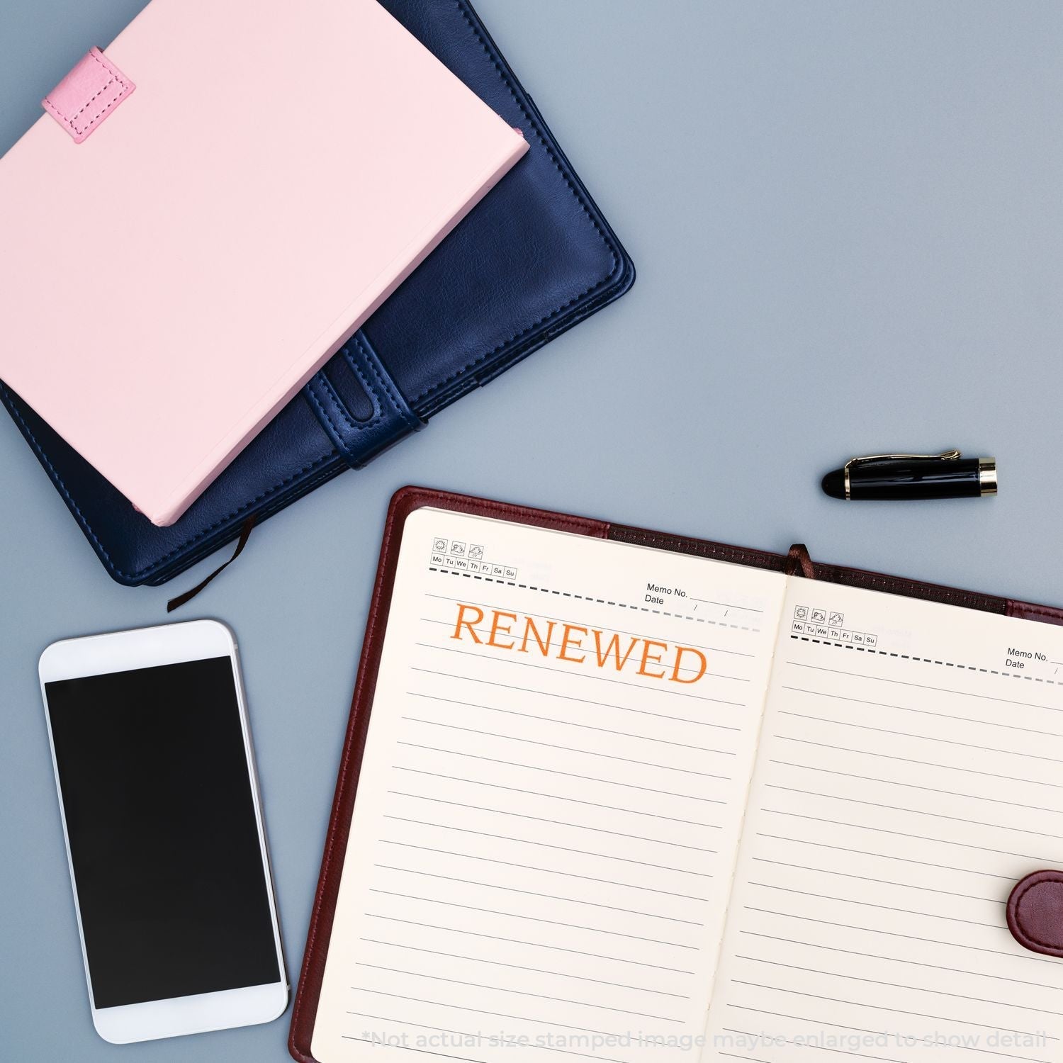 Self Inking Renewed Stamp used on an open notebook, surrounded by a smartphone, pen, and closed notebooks on a gray surface.