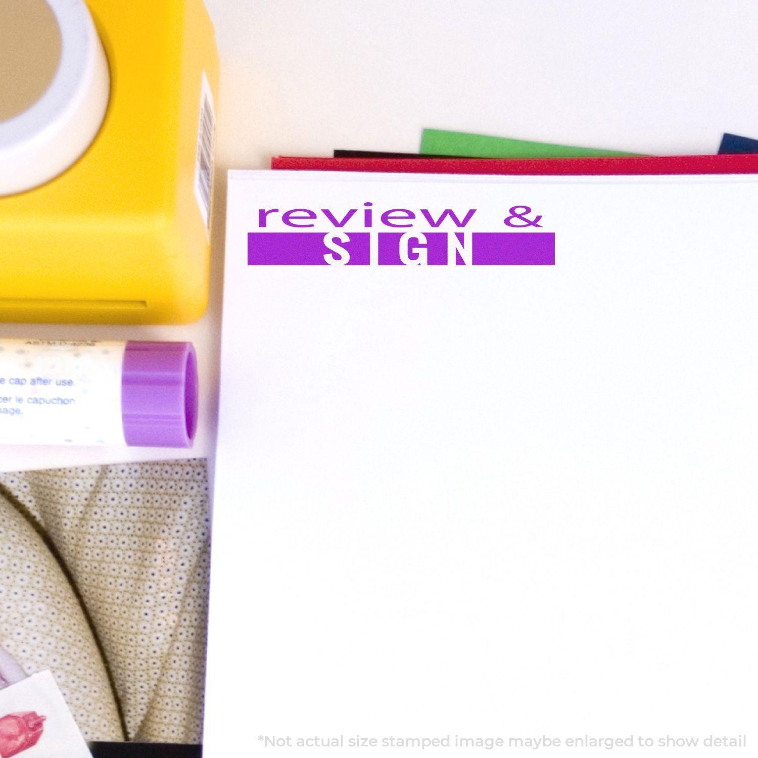 Large Self Inking Review and Sign Stamp in use on a white paper, surrounded by office supplies including a yellow stapler and a purple glue stick.