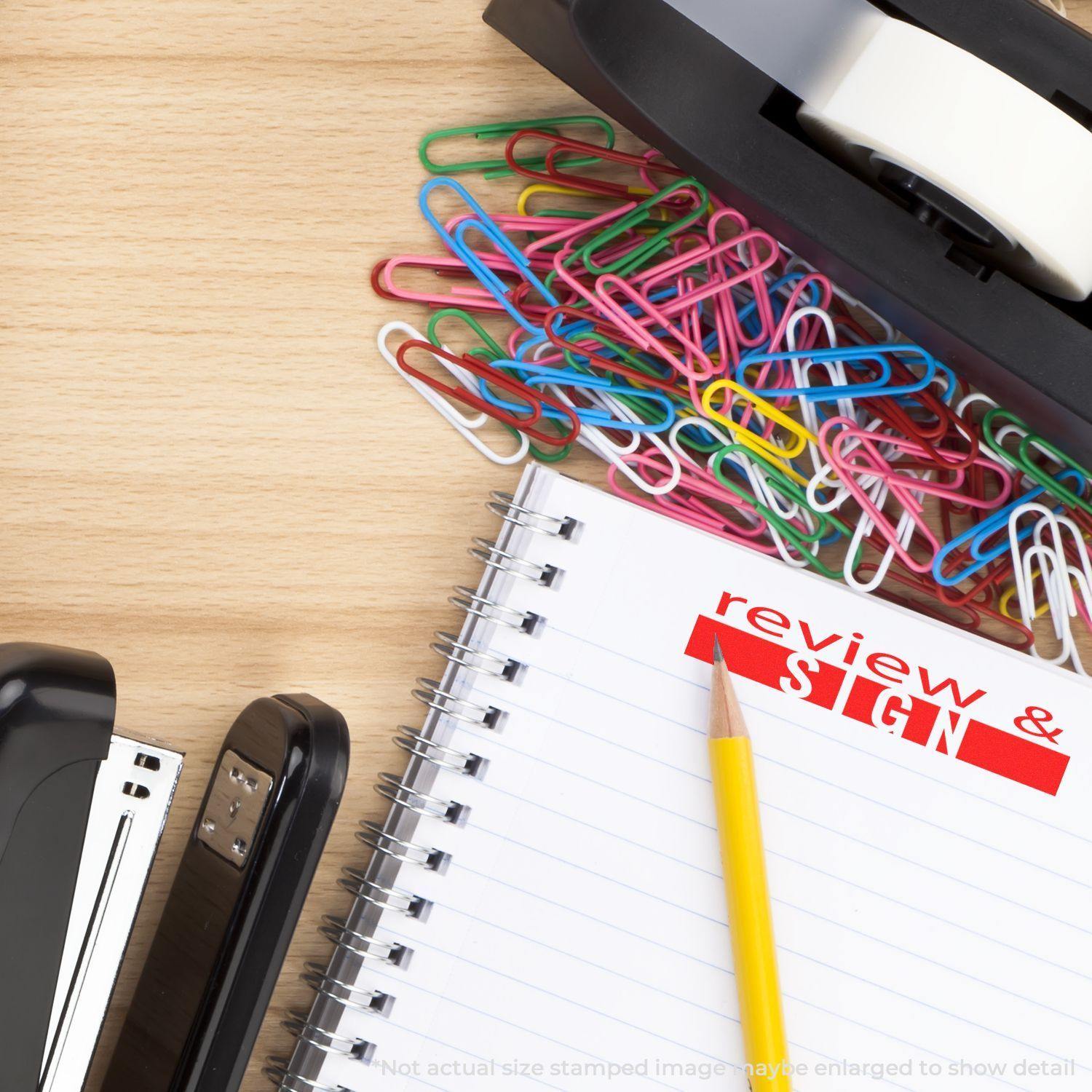 Large Pre-Inked Review and Sign Stamp used on a notebook page with a pencil, stapler, tape dispenser, and colorful paperclips on a wooden desk.