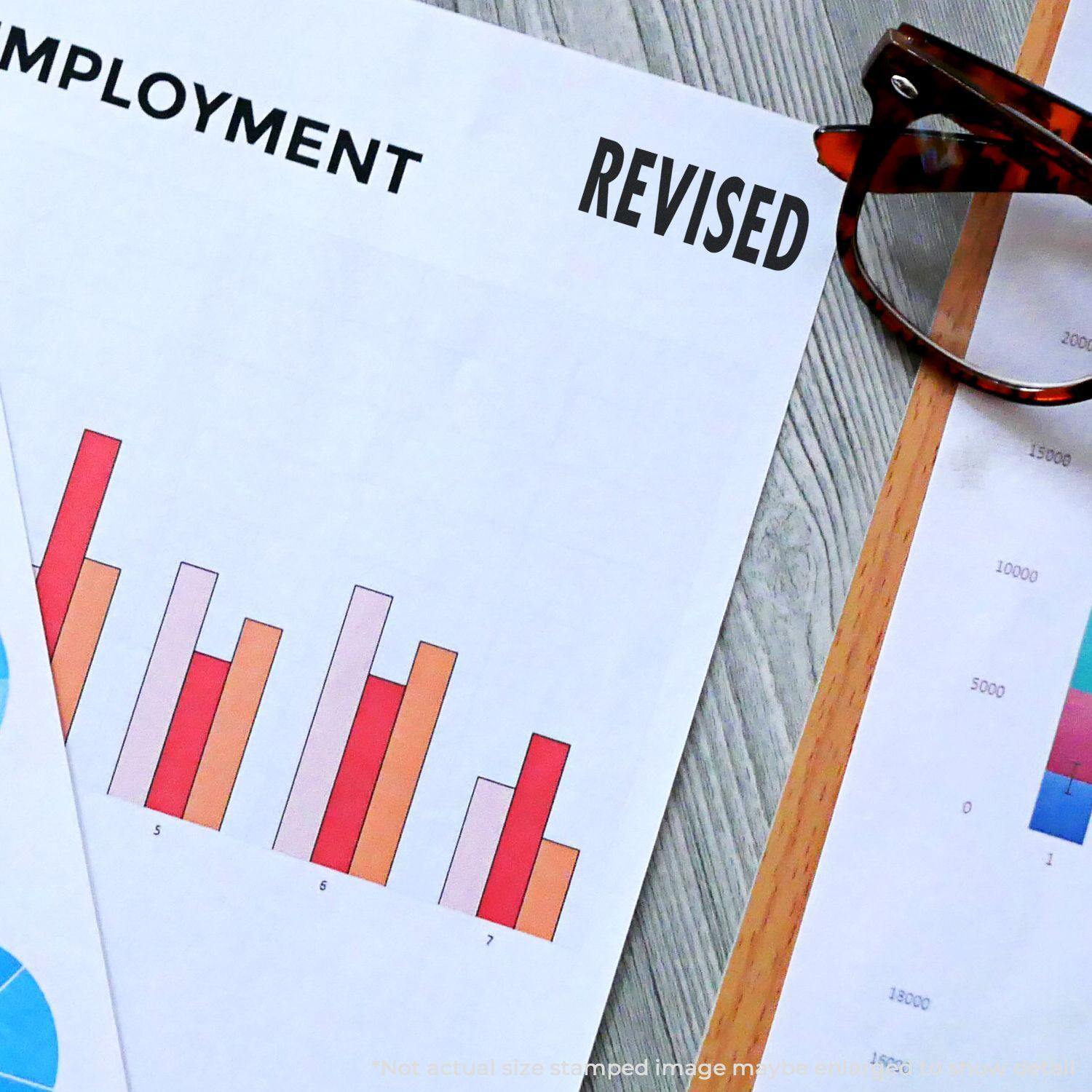 A document with a bar chart is stamped REVISED using the Large Bold Black Revised Rubber Stamp, next to a pair of glasses.