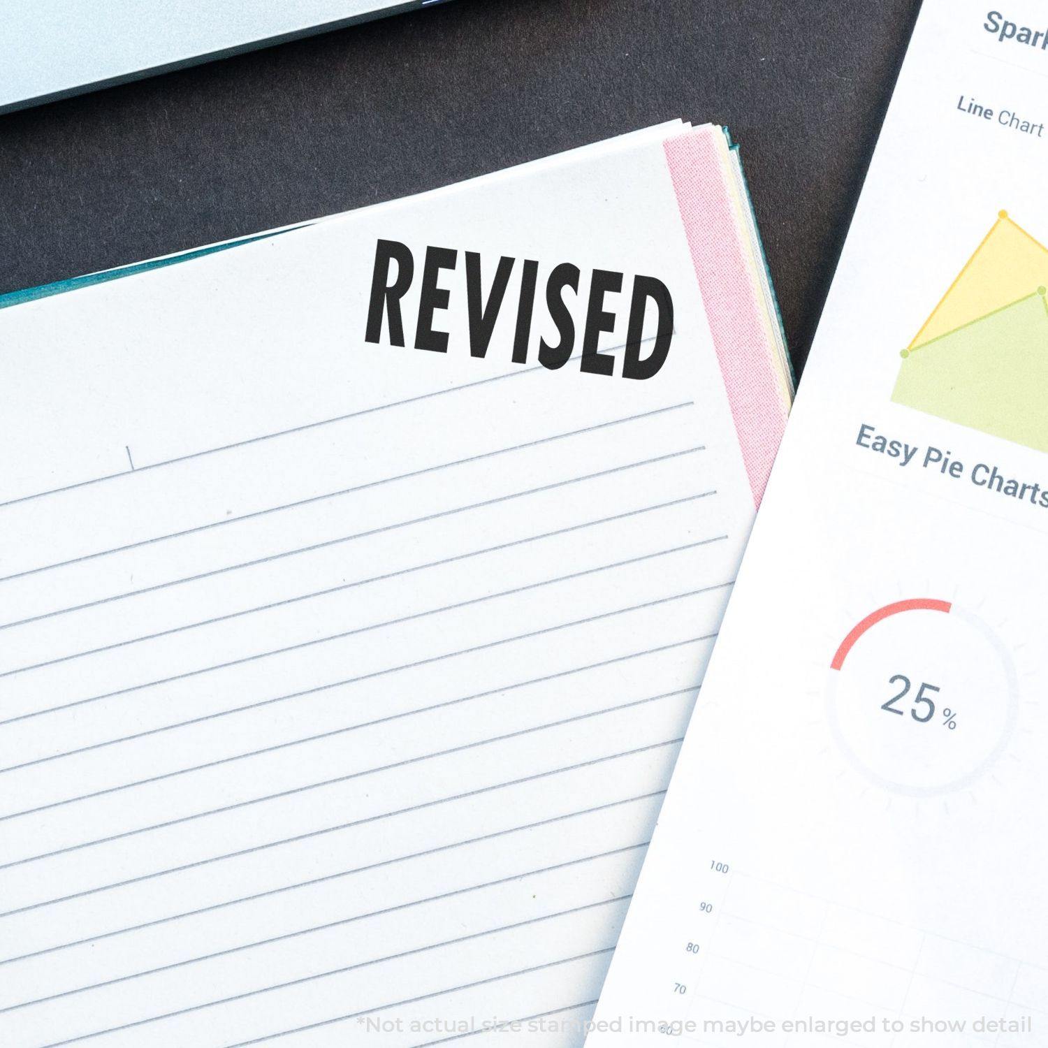 Large Self Inking Bold Revised Stamp marking REVISED on a document, next to a pie chart and a notebook on a desk.