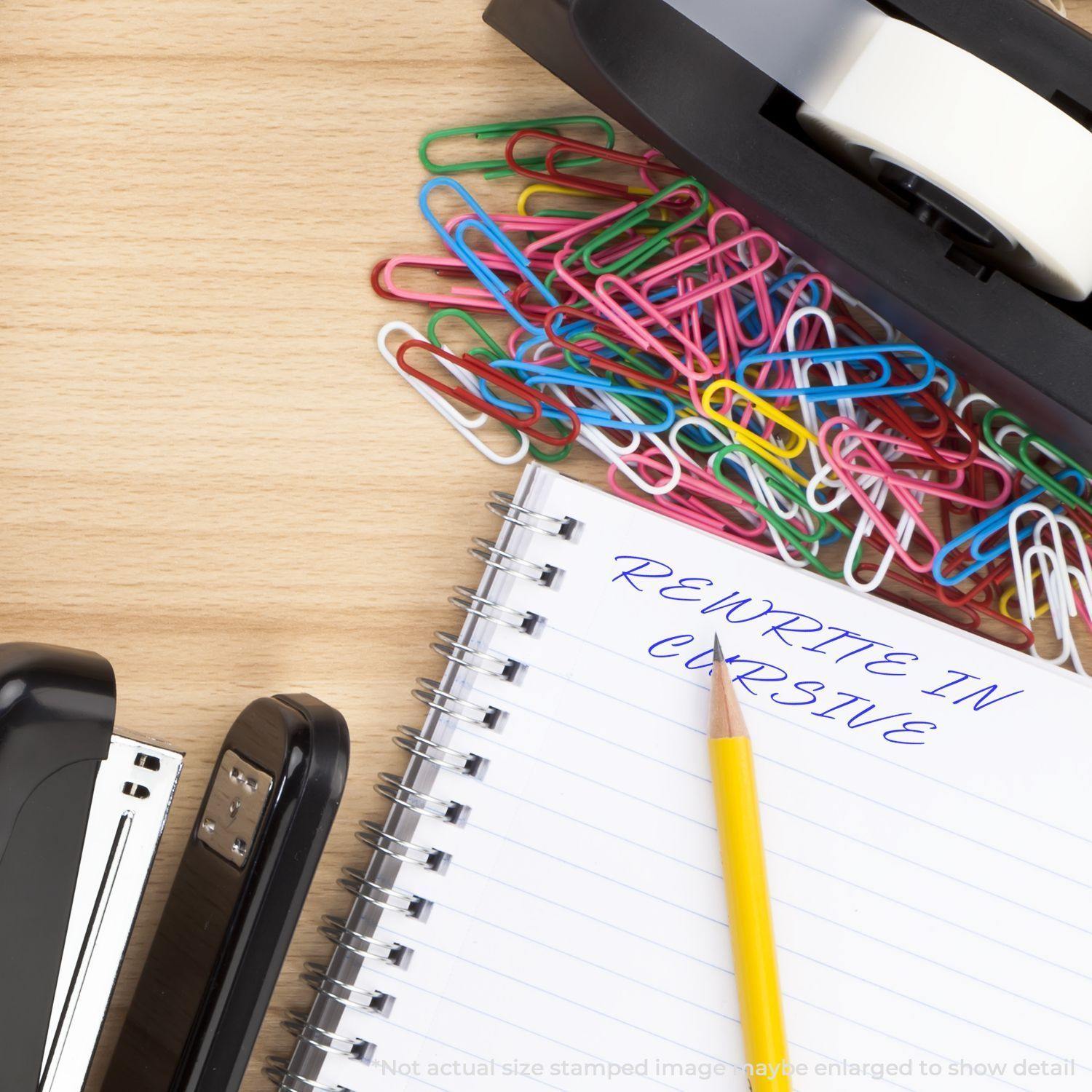 Slim Pre-Inked Rewrite In Cursive Stamp used on a notebook page, surrounded by colorful paperclips, staplers, and a tape dispenser.