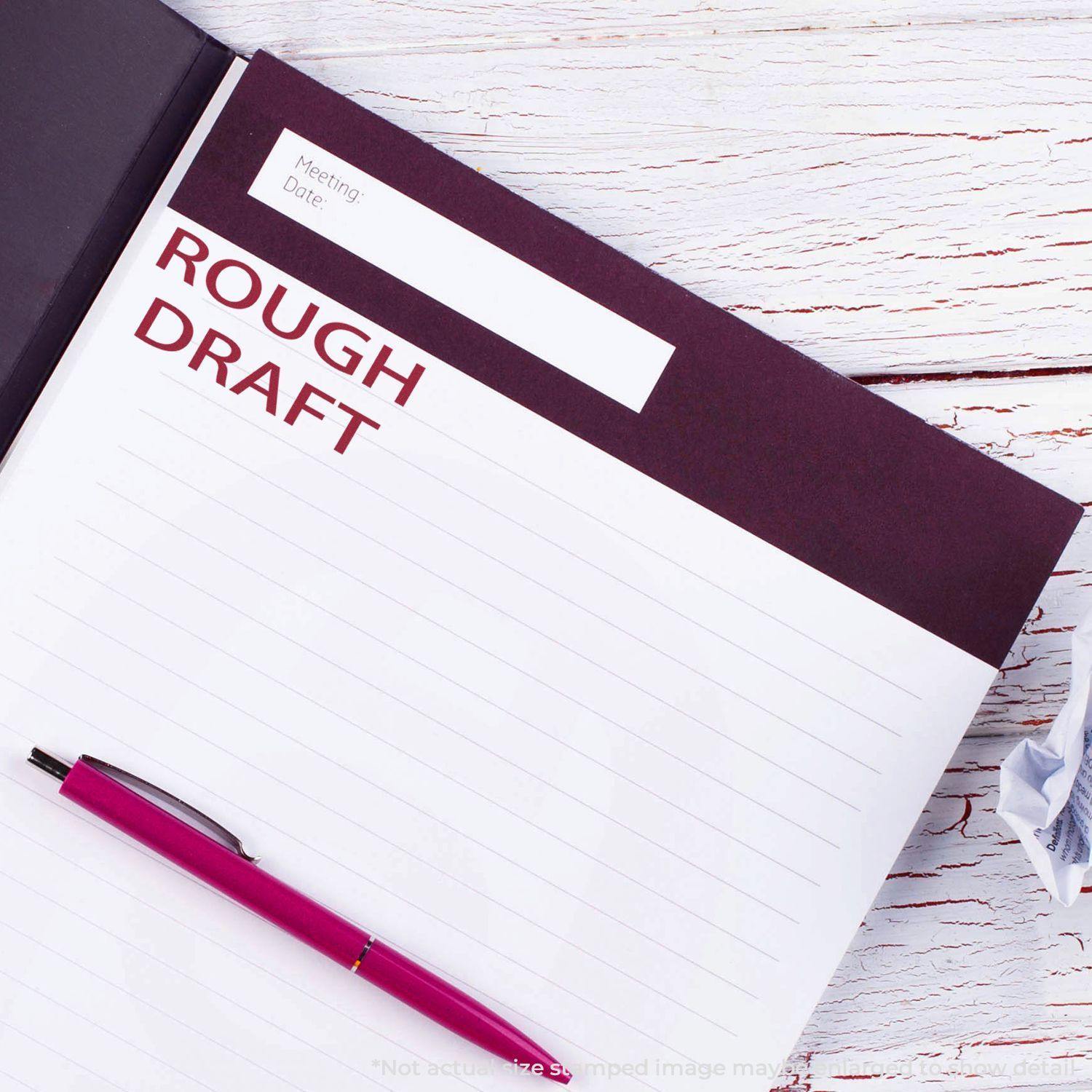 Notebook with 'Rough Draft' rubber stamp in red ink, a pink pen, and a crumpled paper on a white wooden surface.