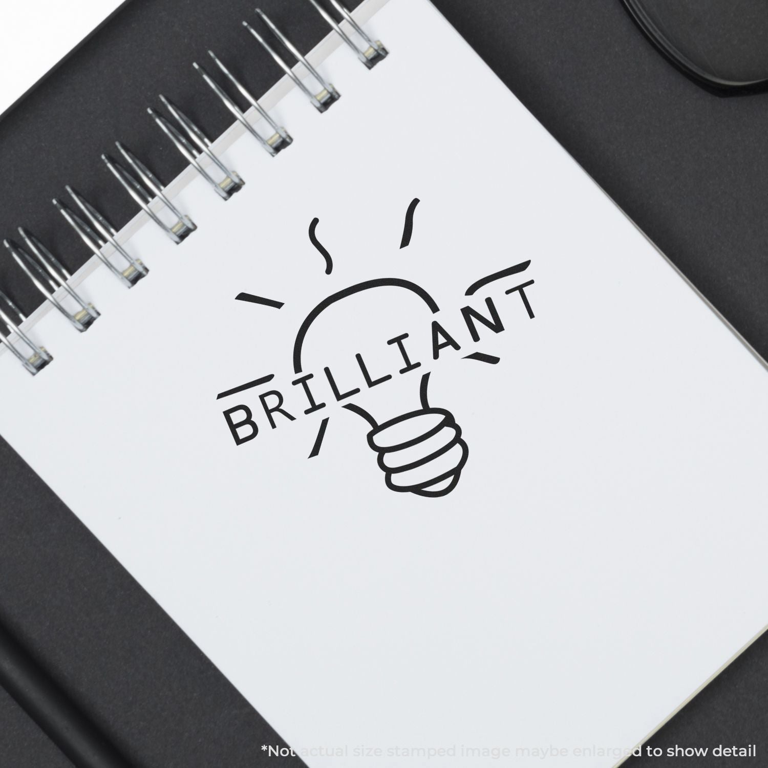 Self Inking Round Brilliant Stamp imprint of a light bulb with the word BRILLIANT on a spiral notebook, placed on a black surface.