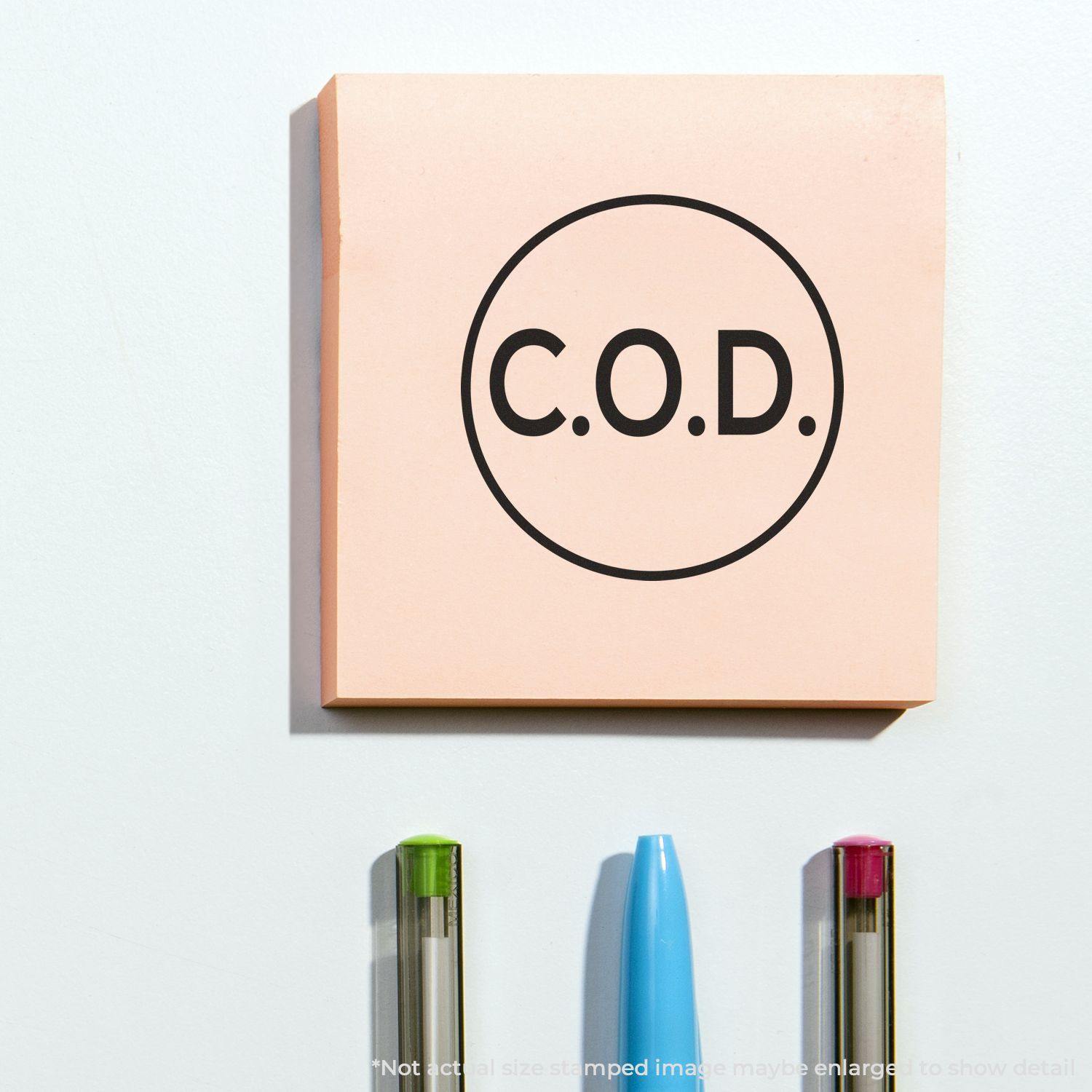 Round COD Rubber Stamp used on a pink sticky note with three pens below it. The stamp displays "C.O.D." inside a circle.