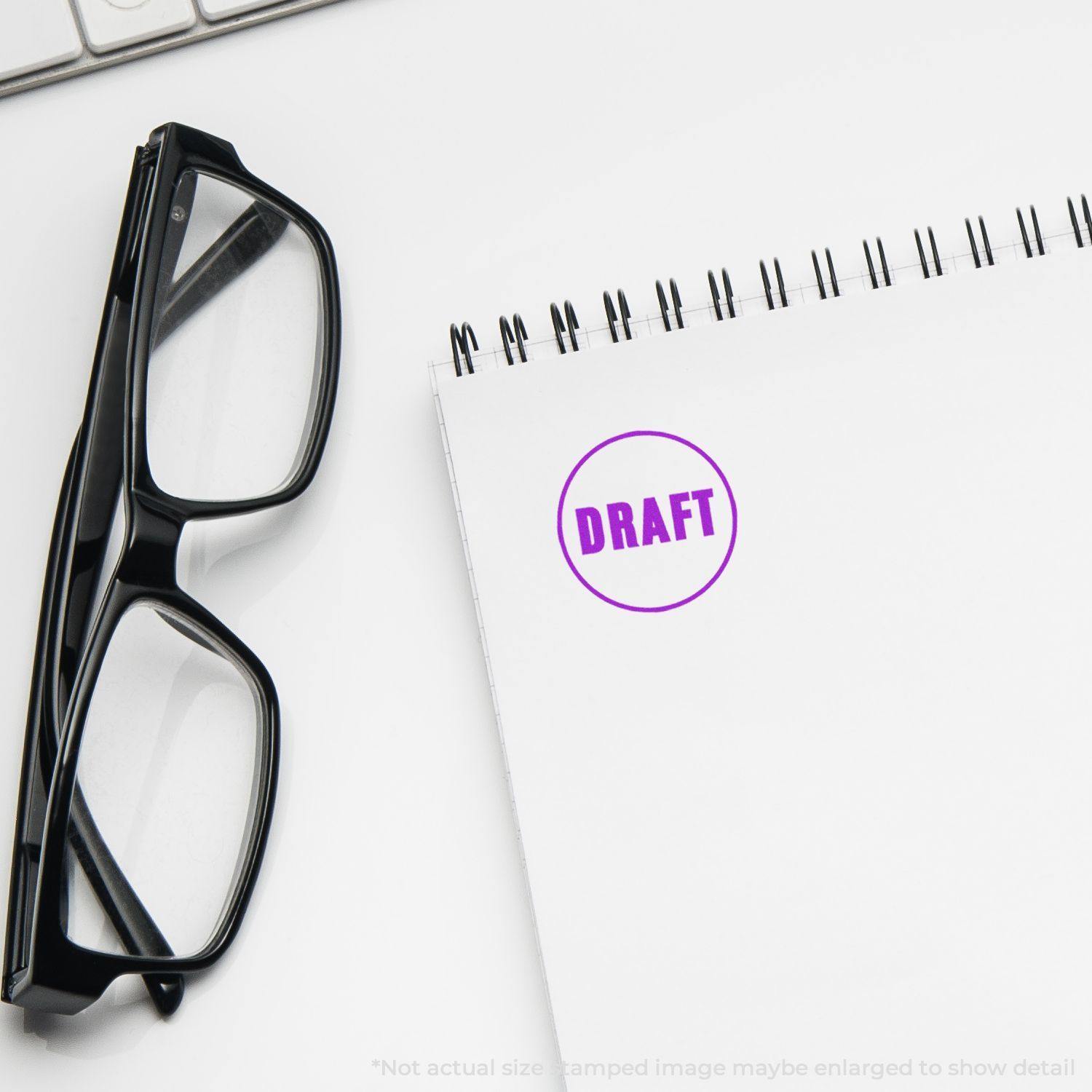 Round draft rubber stamp in purple ink on a white notepad, with black-rimmed glasses and a keyboard in the background.