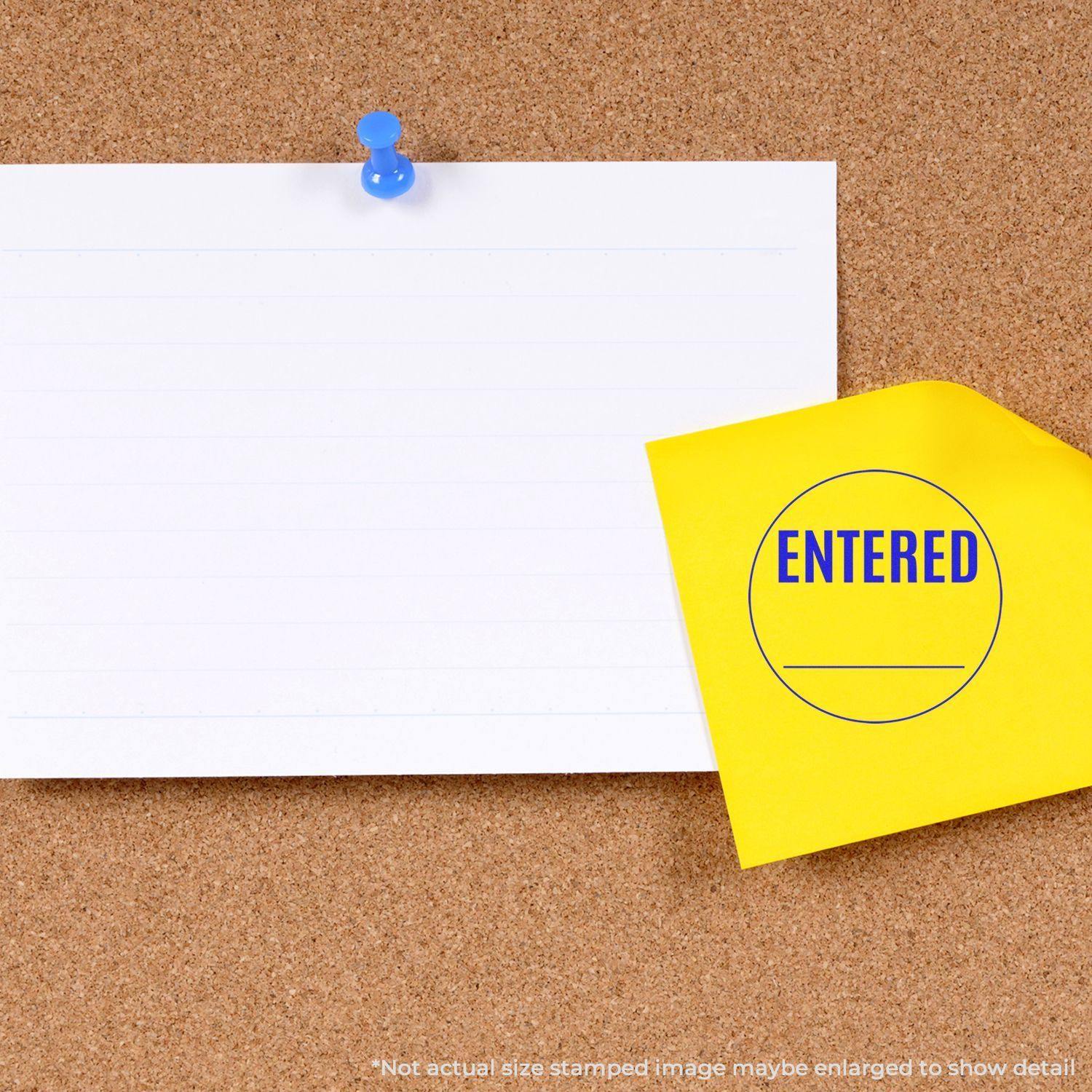 A round entered rubber stamp on a yellow sticky note pinned to a corkboard, next to a blank white paper with a blue pushpin.