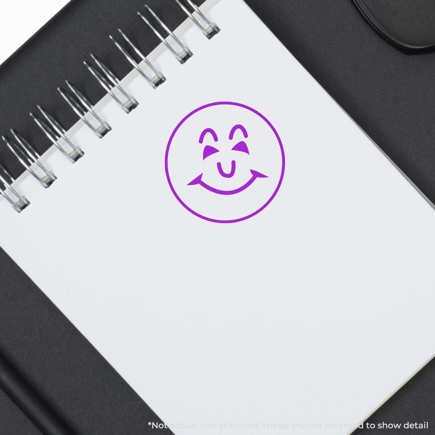 Self Inking Round Happy Face Stamp in purple ink on a white spiral notebook, placed on a black surface.