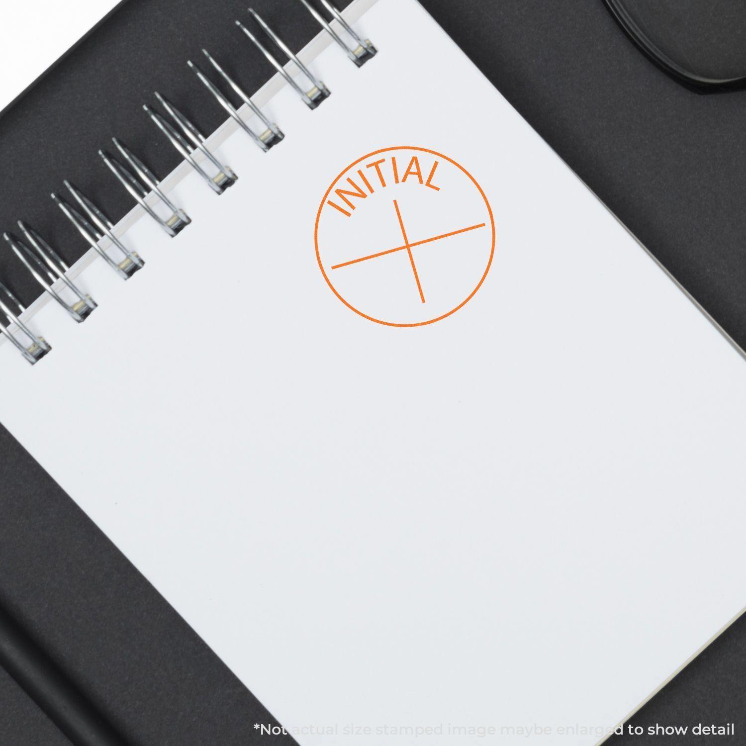 Round Initial Rubber Stamp in orange ink on a white notepad with a spiral binding, placed on a black surface.