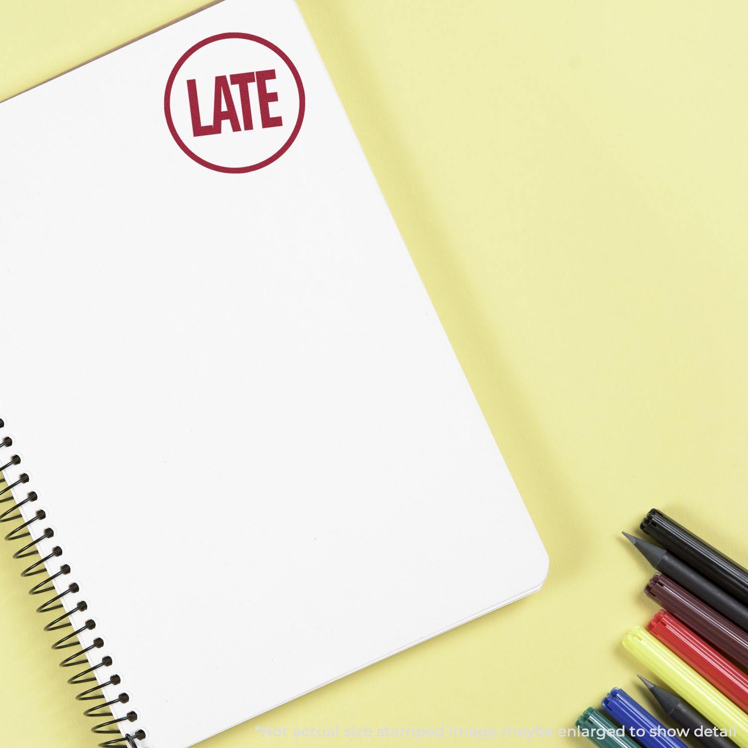 A round LATE rubber stamp mark on a white spiral notebook, with colorful pens and markers on a yellow background.