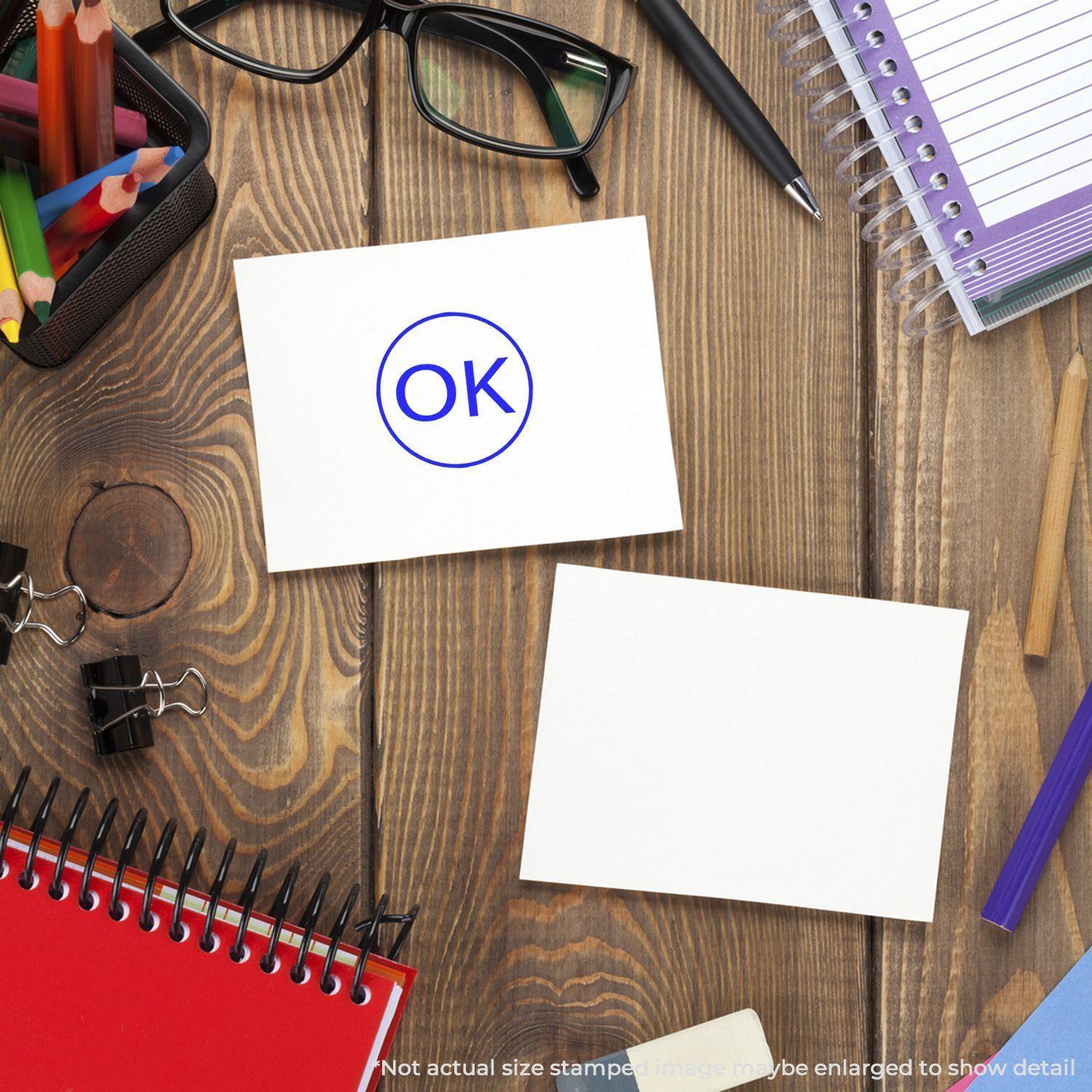 A Round OK Rubber Stamp on a white card, surrounded by office supplies like notebooks, glasses, and pens on a wooden desk.