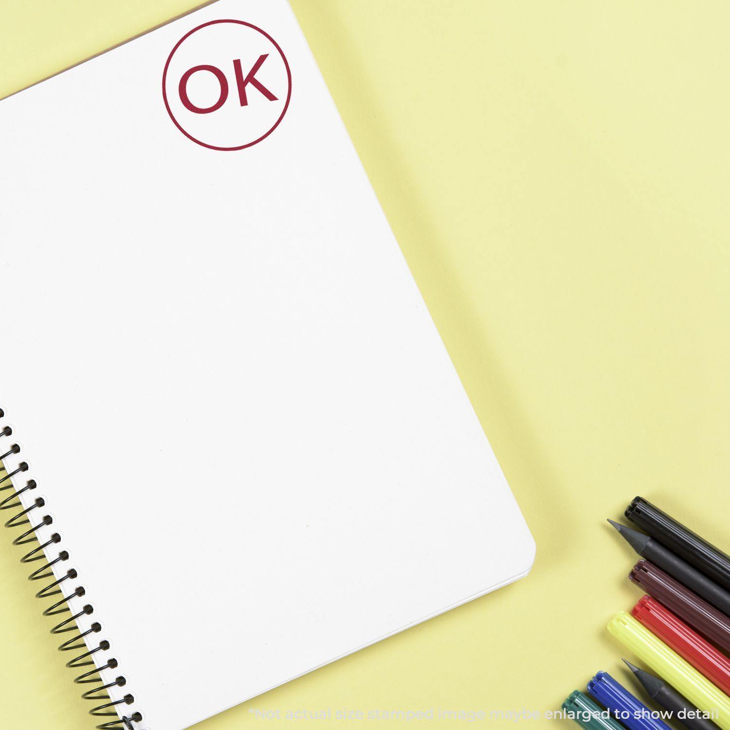 Round OK rubber stamp on a blank spiral notebook page with colored pens on a yellow background.