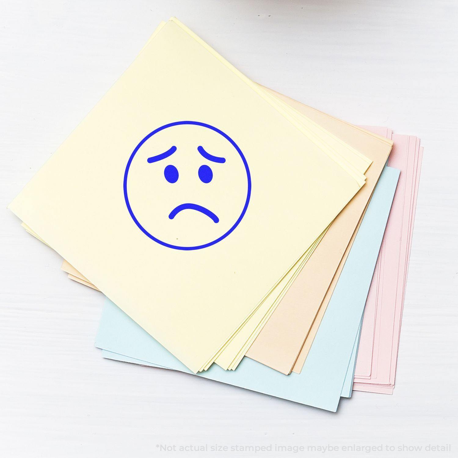 Round Sad Face Rubber Stamp on a stack of pastel-colored papers, showing a blue sad face on the top yellow sheet.