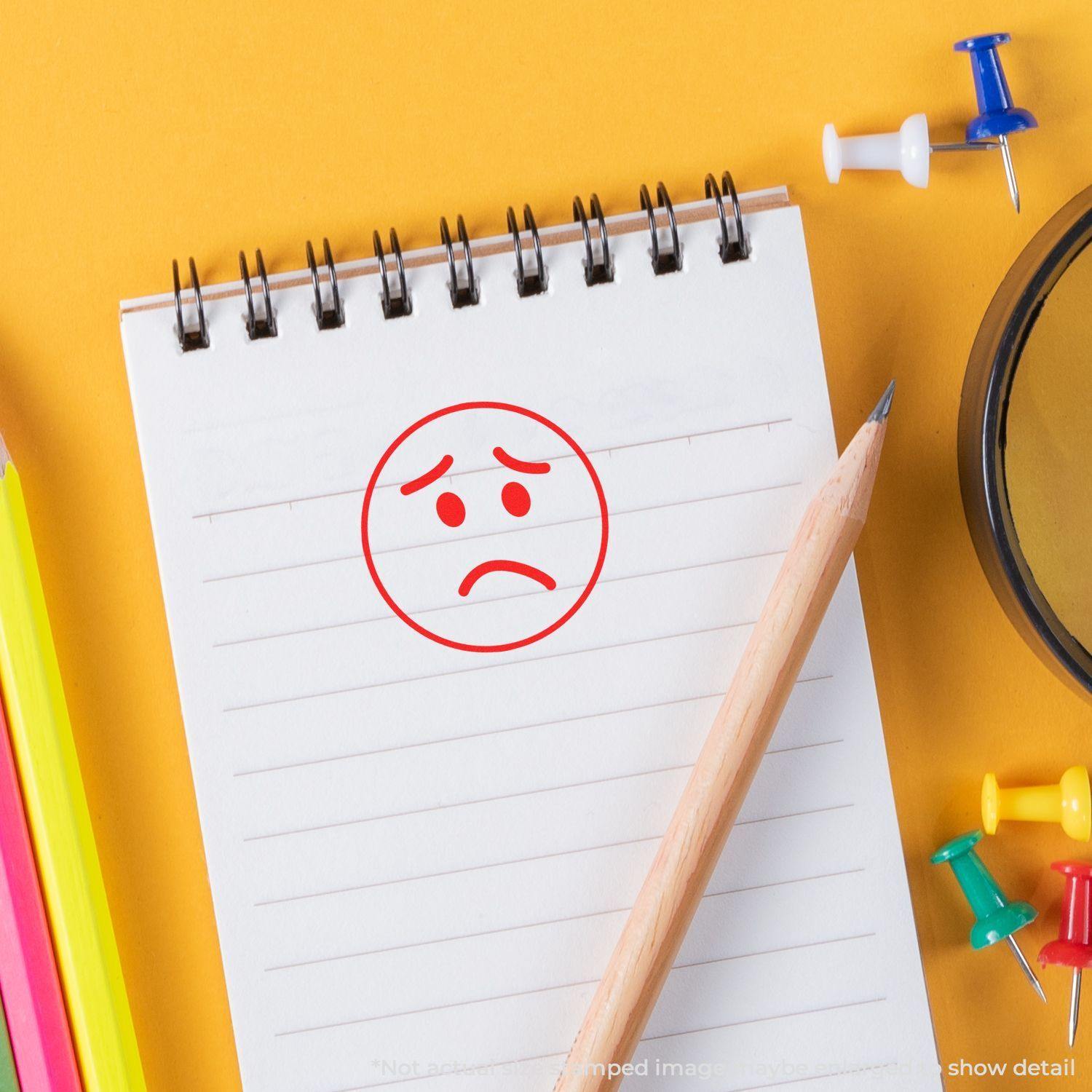 Round Sad Face Rubber Stamp on a notepad with a pencil, surrounded by colorful push pins and stationery on a yellow background.